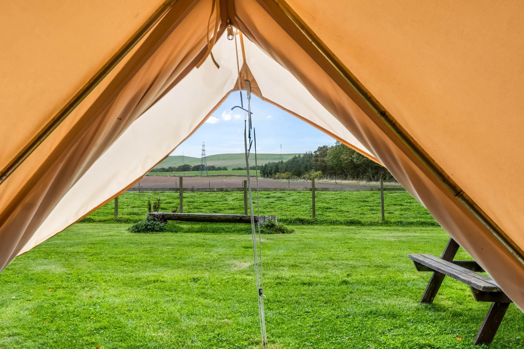 View through the open flap of a tent showing a green grassy field, a wooden picnic table, a fence, and rolling hills in the distance under a partly cloudy sky.