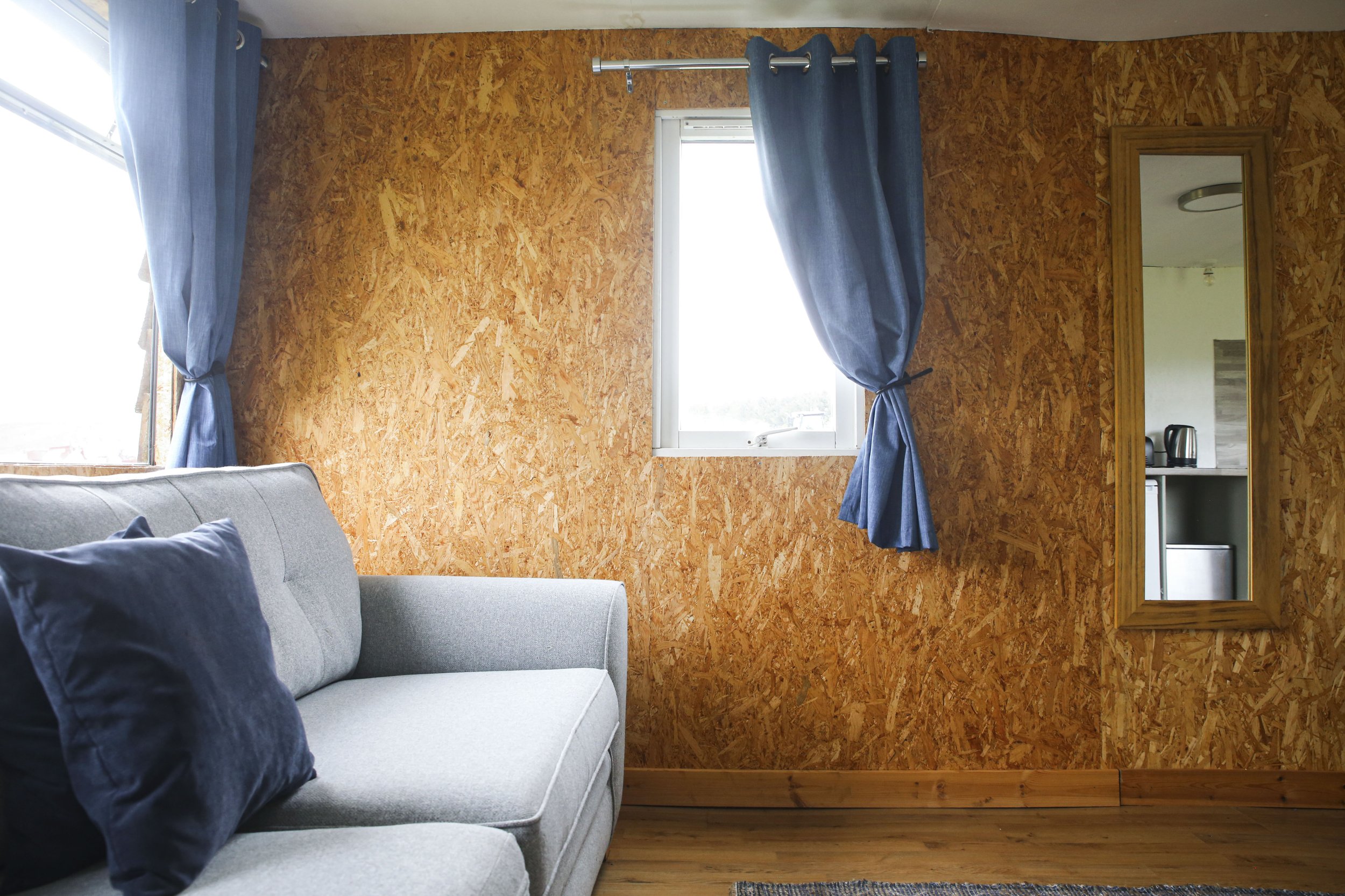 A living room with a gray sofa with navy pillows, a wooden wall, a window with blue curtains, a full-length mirror with a wooden frame, and a partial view of a kitchenette with a kettle and a mini fridge.