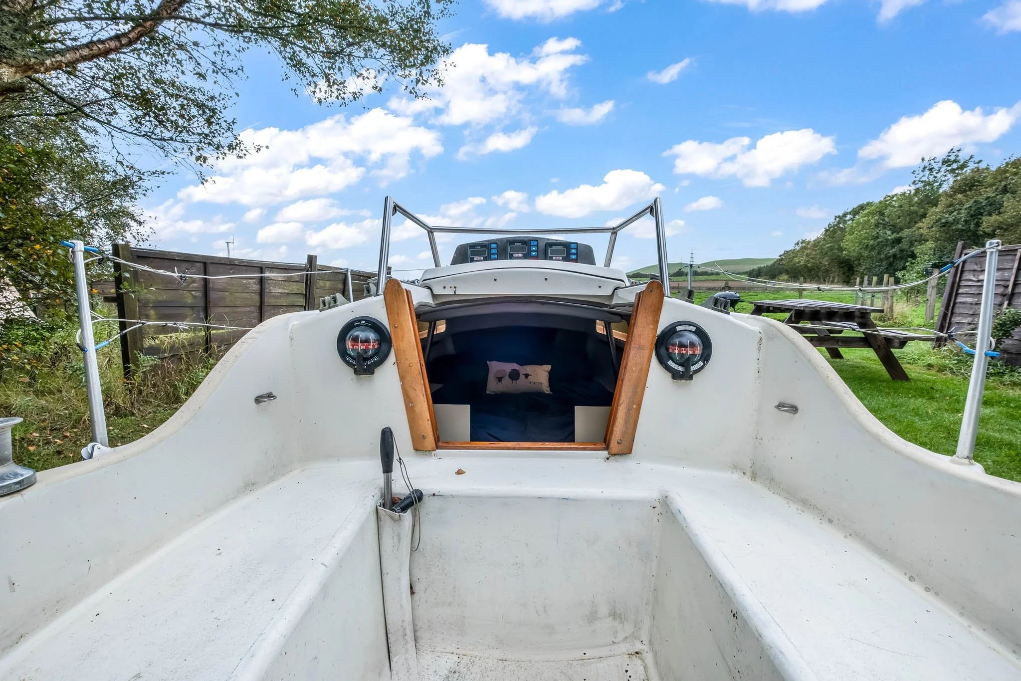 Front view of an empty boat in a grassy outdoor area with trees, fence, and picnic table, under a partly cloudy sky.