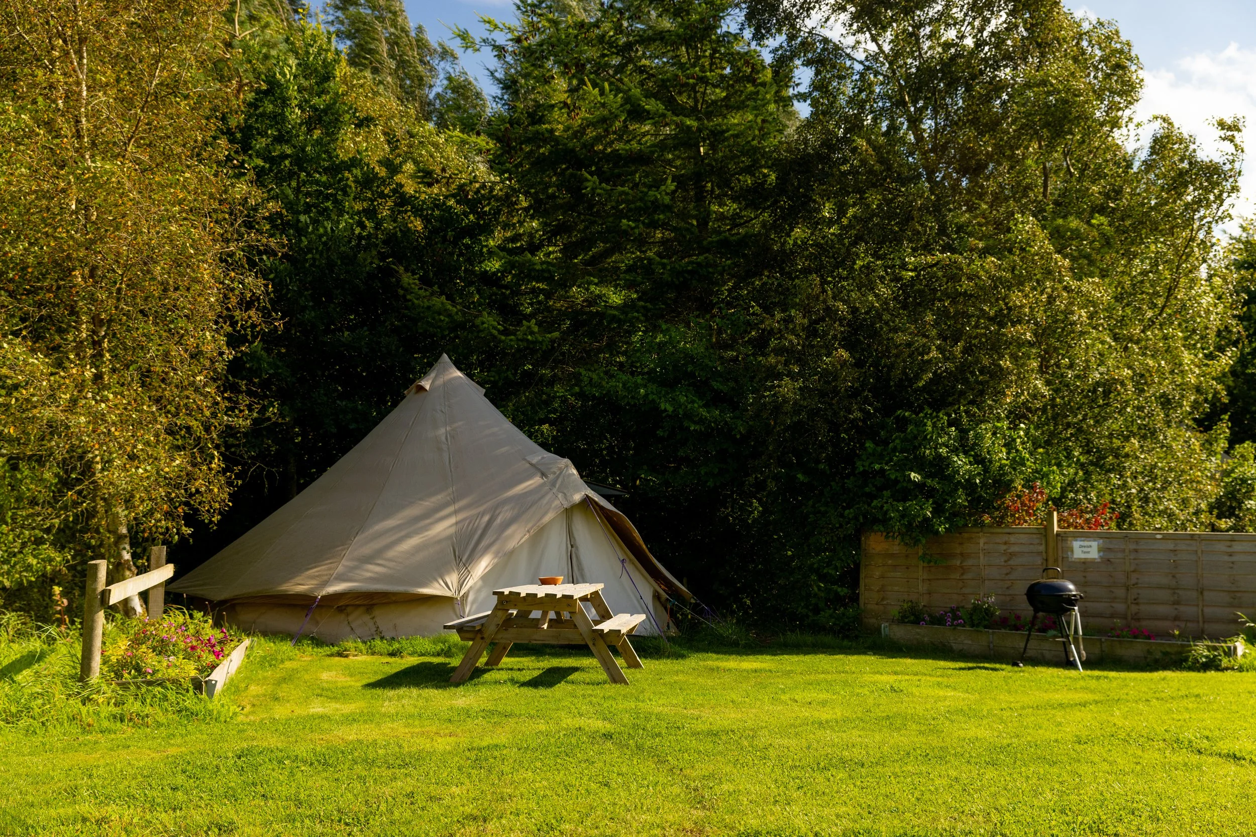A large beige canvas tent set up on a grassy lawn surrounded by green trees. There is a wooden picnic table with a bowl on top and a small barbecue grill on the right, next to a wooden fence.