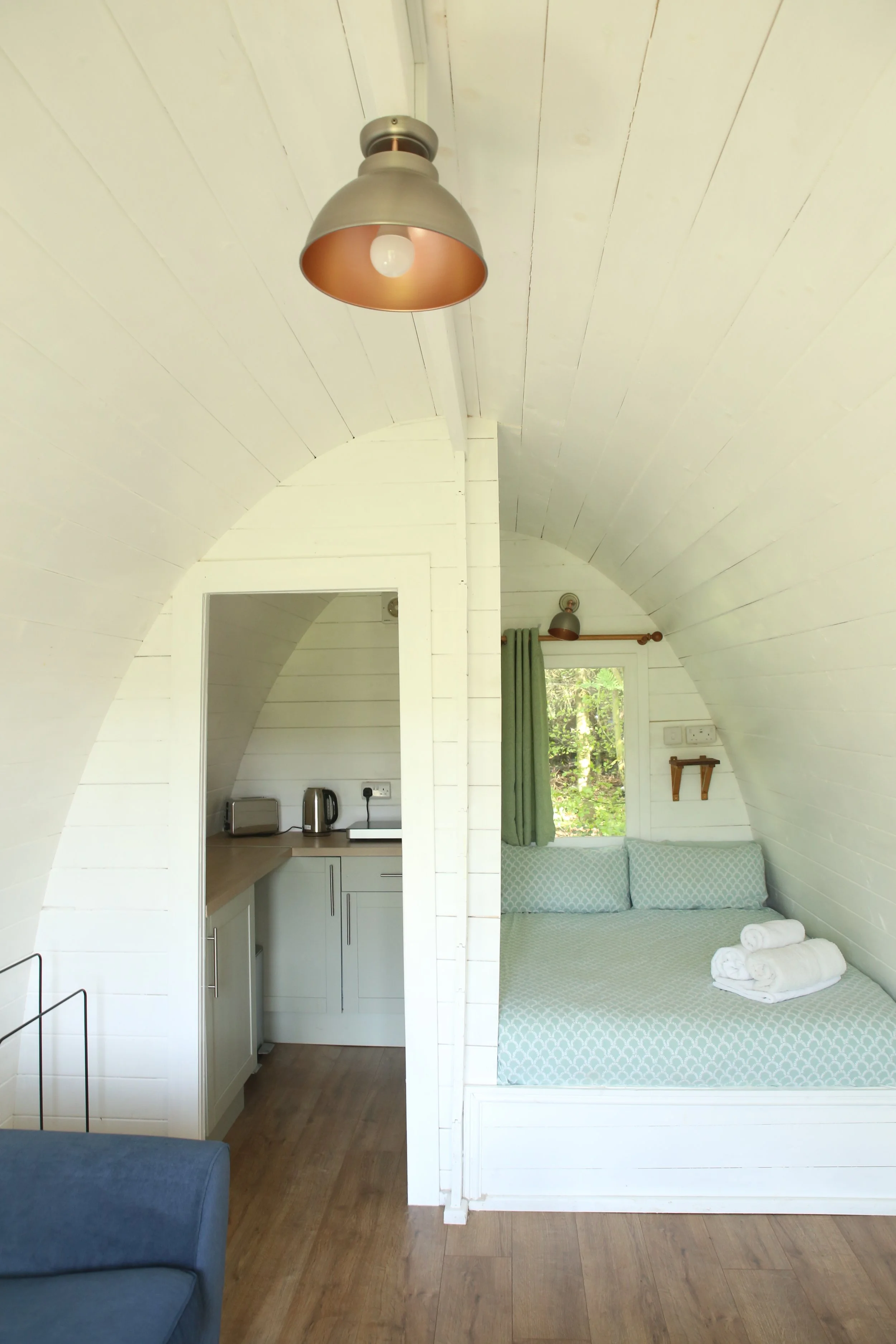 Interior of a small cozy cottage with a white wooden arched ceiling, featuring a bed with neatly folded towels, windows with green curtains, a small kitchen area with appliances, and a wooden floor.