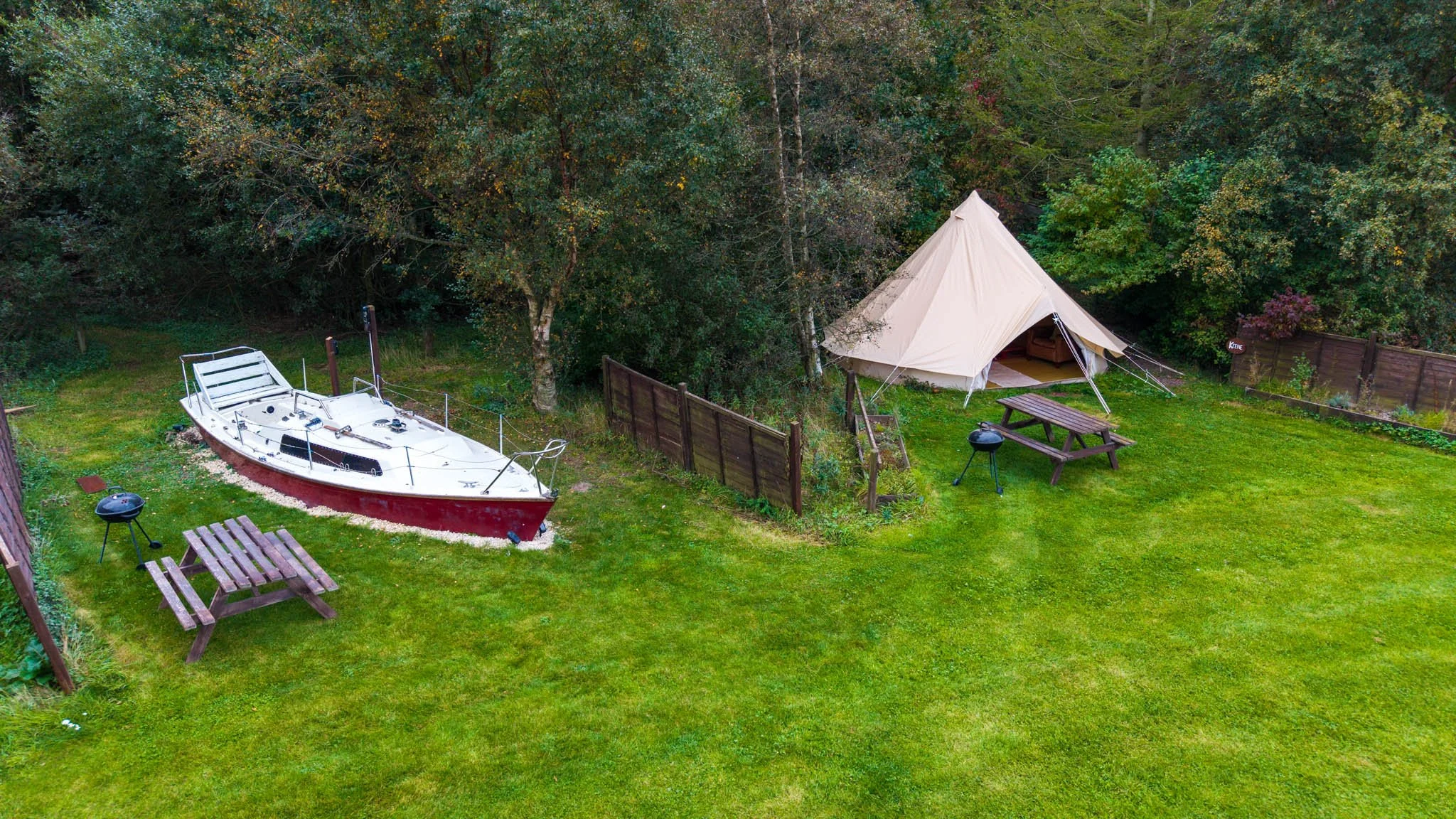A backyard with a boat on a gravel bed, a canvas tent, picnic tables, small grills, and green grass, surrounded by trees and a wooden fence.