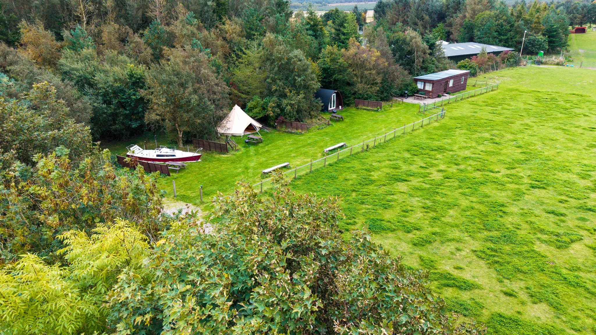 Aerial view of a grassy field with a fenced area and trees in the background. There are two small tents, a boat, picnic tables, benches, and small buildings near the tree line.