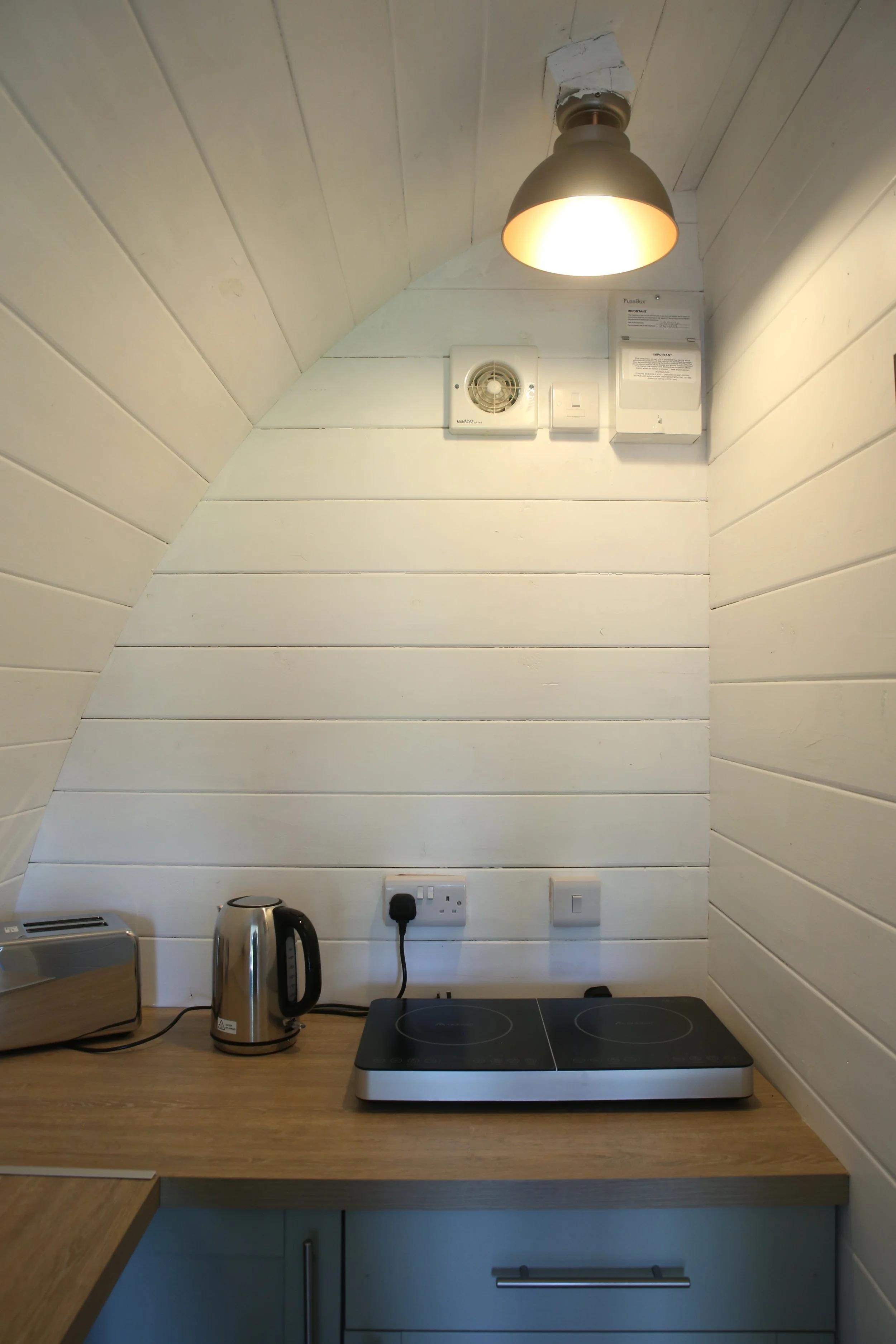 Kitchen corner with a small wood countertop, electric kettle, toaster, two electrical outlets, and a Double induction cooktop under a wall-mounted lamp. The wall is painted white with horizontal wood paneling.