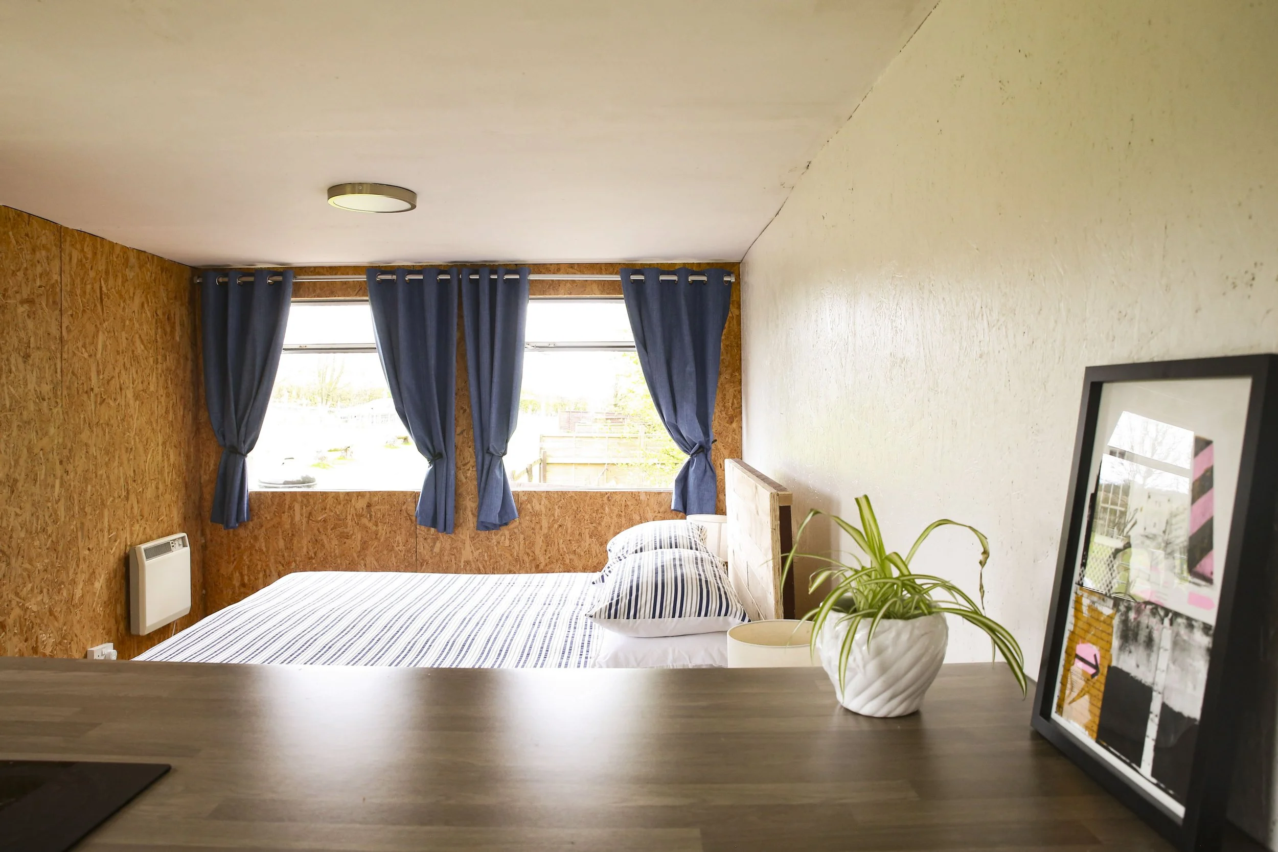 A bedroom with a bed featuring striped bedding, a potted plant on a wooden countertop, a framed picture, and blue curtains over large windows letting in natural light, with a wall heater on the left.