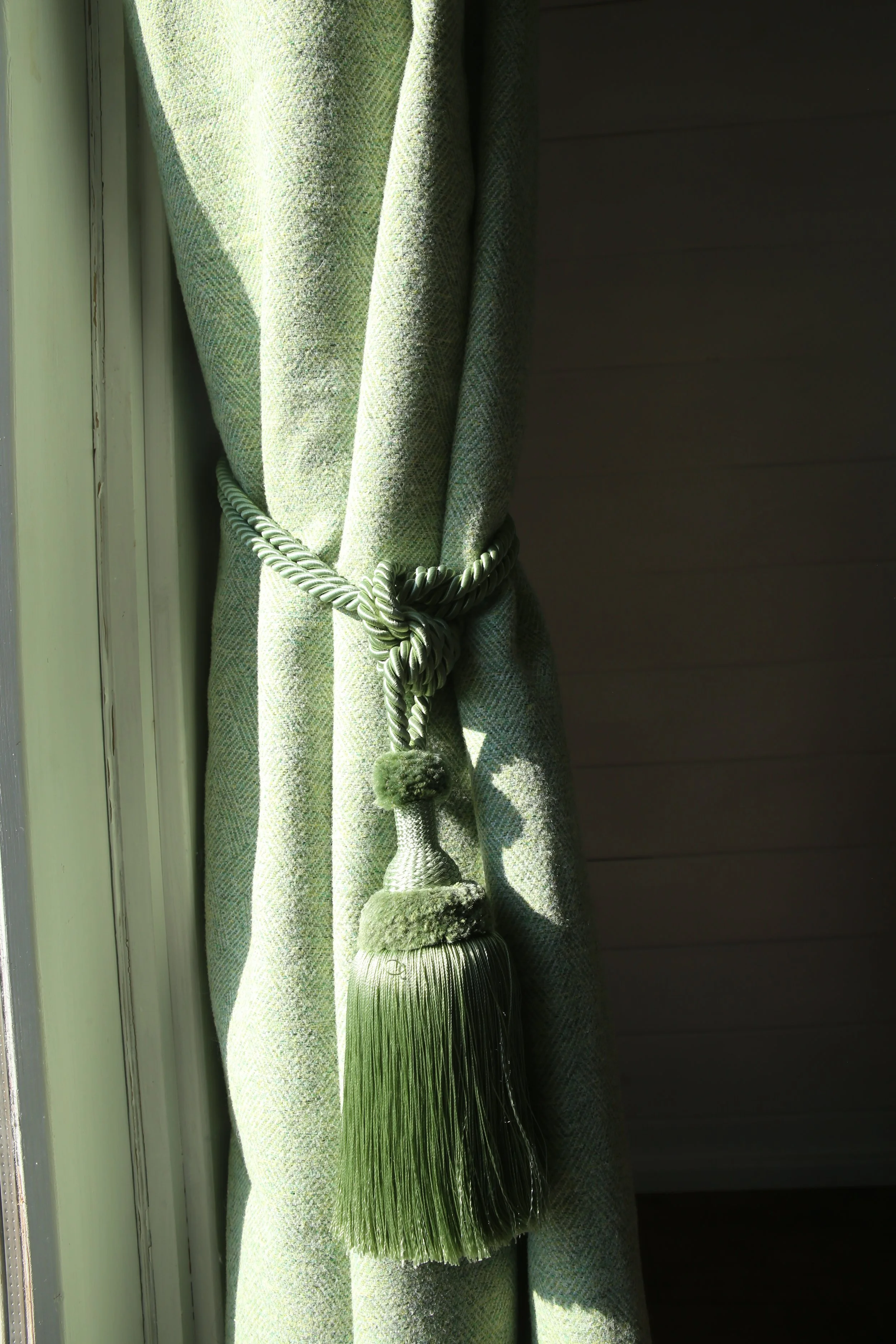 Green curtain tied back with a decorative tasseled tie, sunlight shining on fabric.