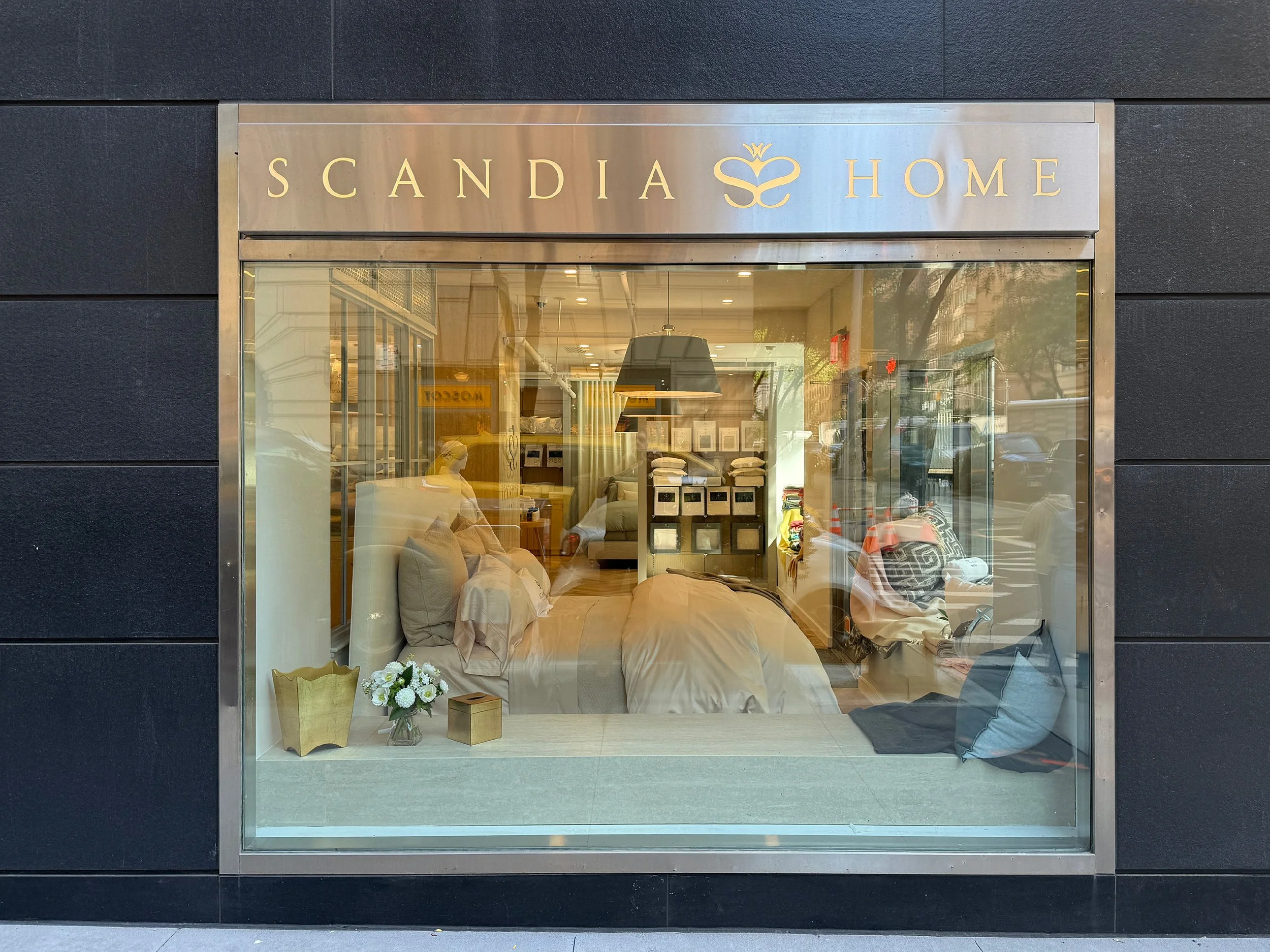 Storefront window displaying a furniture or bedding store called 'Scandia Home' with interior beds, pillows, and decor visible through the glass, with reflections of street outside.