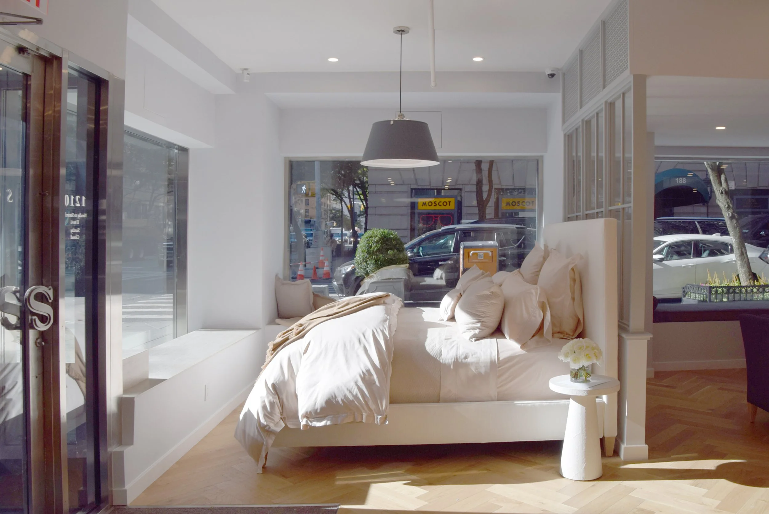 Bright interior of a modern café with a white bed serving as a seating area, decorated with white pillows and a blanket. Large front windows reveal parked cars and street scene outside, with a black pendant light hanging from the ceiling.