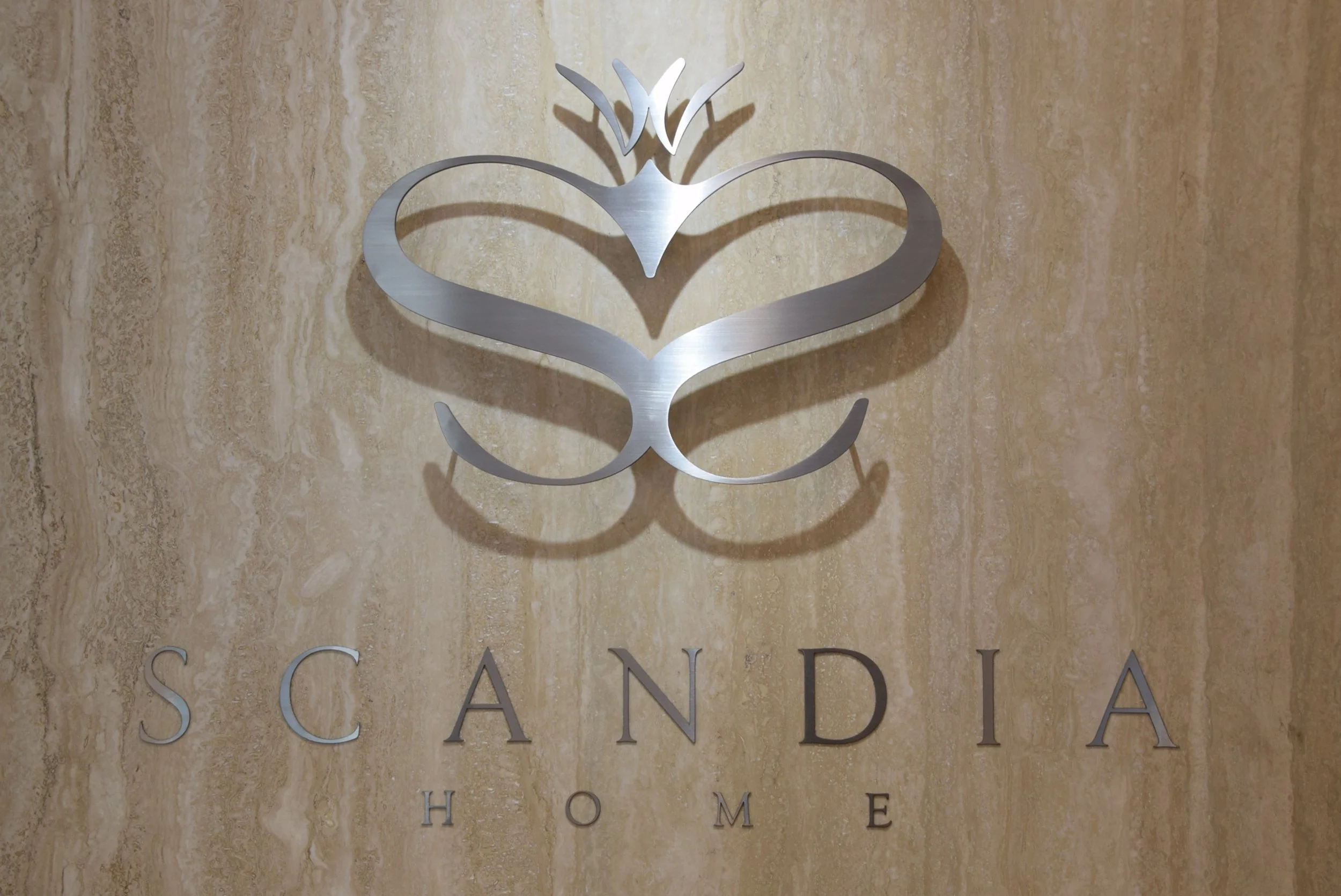 A metallic logo of a heart and crown design above the word 'Scandia' in large letters, with 'Home' written below, on a beige travertine wall.