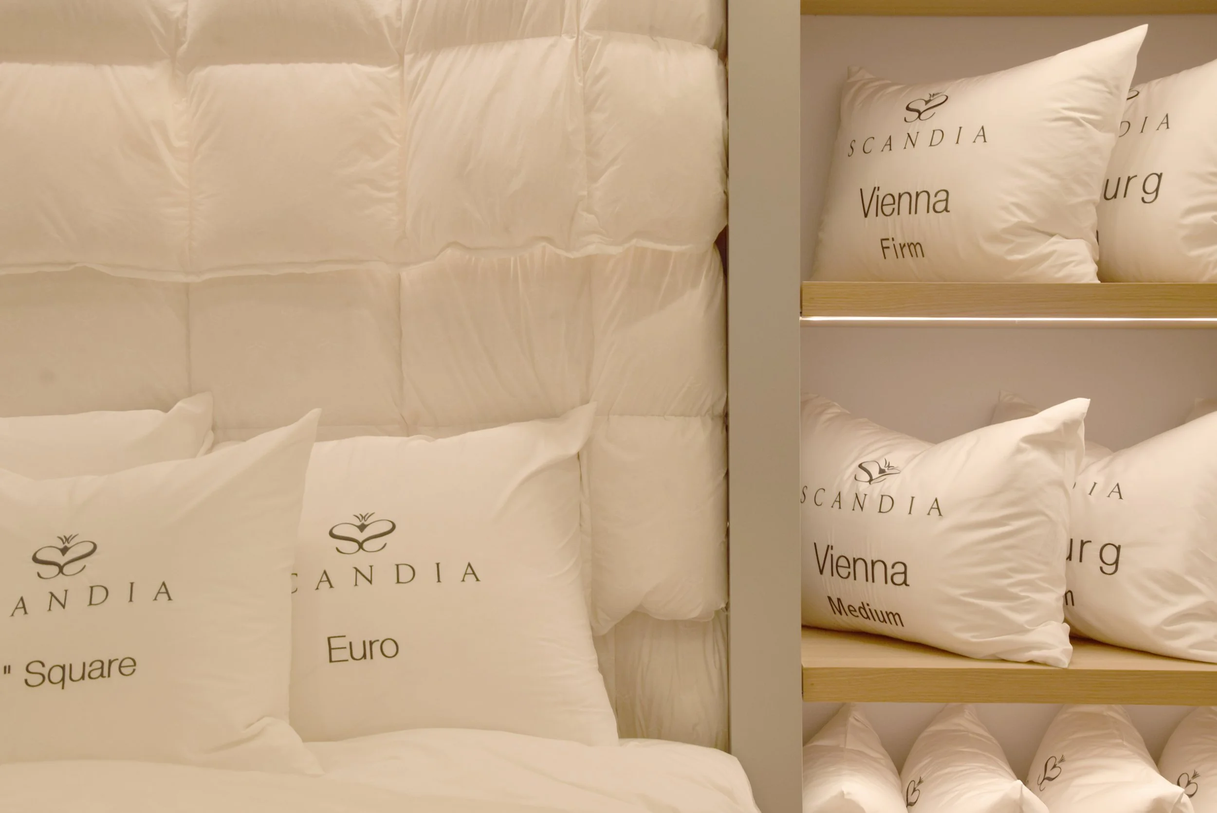 Display of white pillows with black text and logos, arranged on a down bedding display inside store shelves. The pillows are branded 'Scandia' and labeled with different names like 'Euro', 'Square', 'Vienna', 'Firm', and 'Medium'.