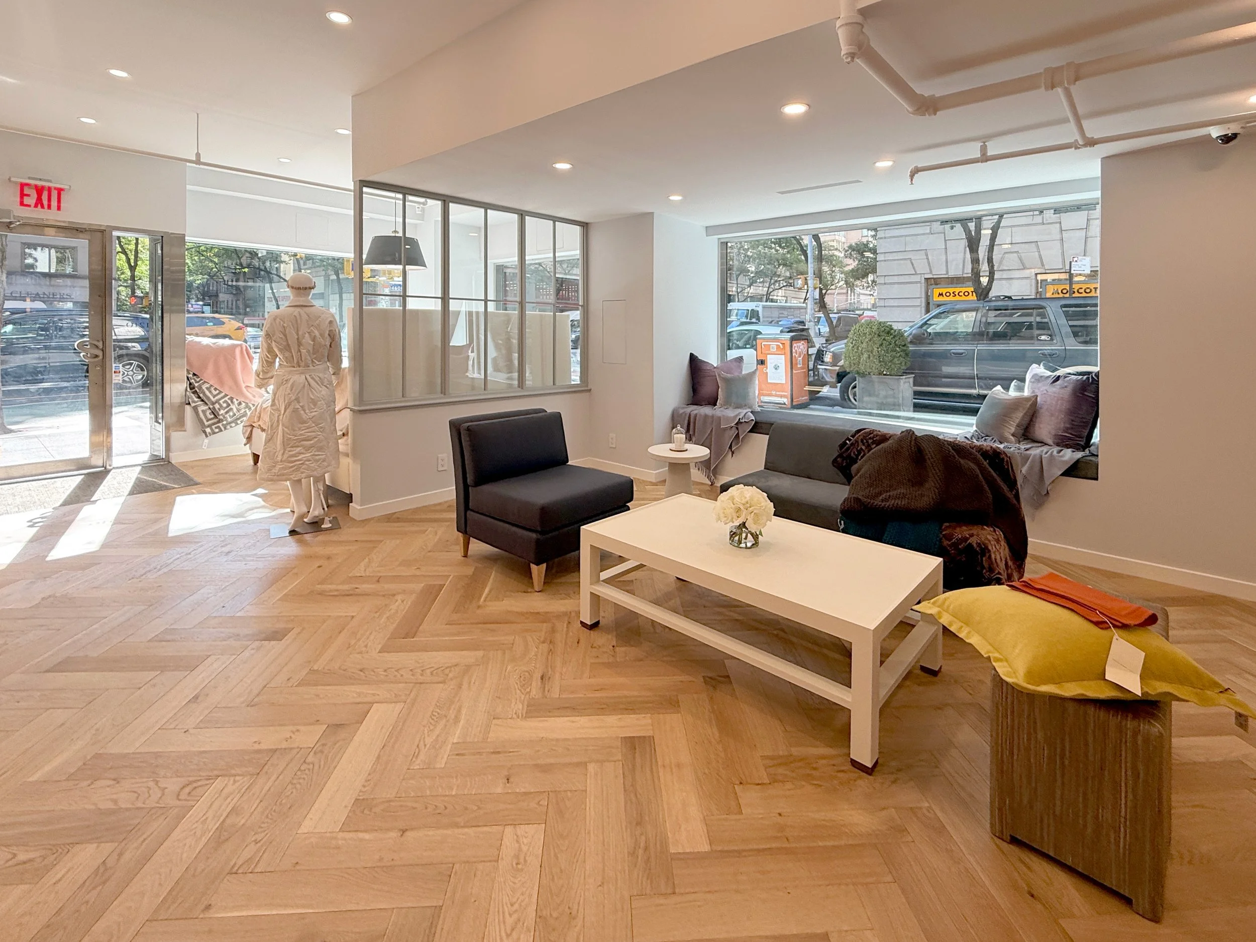 Interior of a modern store or waiting area with large windows, a front door, a mannequin dressed in a bathrobe, a black armchair, a gray sofa, a white coffee table, and various cushions and decor. Outside, cars and a city street are visible.