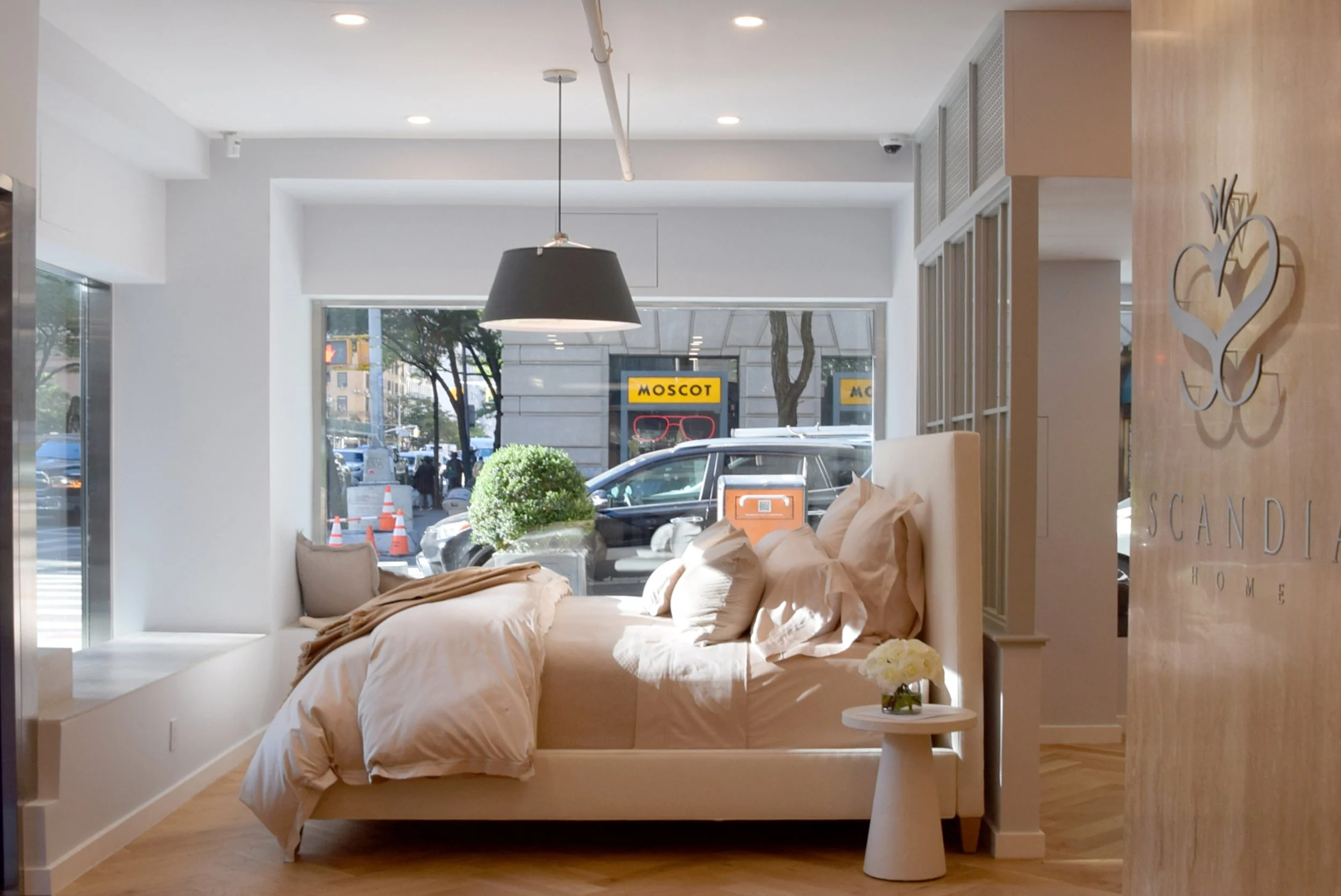 Interior of a bed store showroom with a neatly made bed, beige bedding, and a small white side table with a vase of white flowers