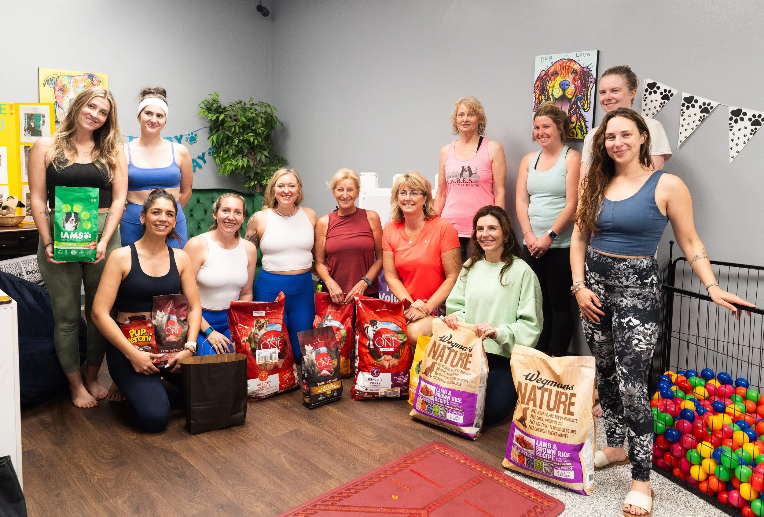 Five women practicing yoga on a beach, standing on colorful yoga mats in tree pose with arms raised, overlooking the ocean with a mountainous coastline in the background.
