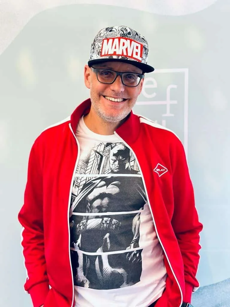 Ein Mann trägt eine Marvel-Cap, eine Brille und ein T-Shirt mit Batman-Comic-Motiv, während er lächelt.