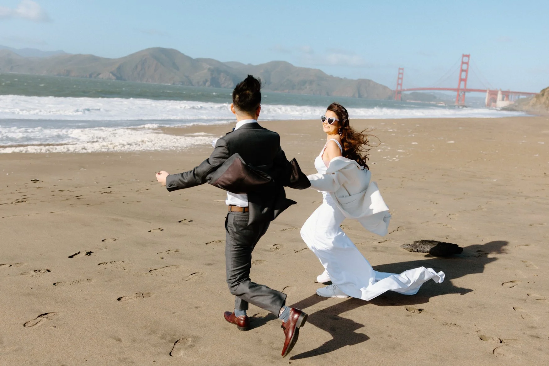 Candid wedding photo of Carla and Jonathan Running in Baker Beach with the Golden Gate Bridge in the Backdrop