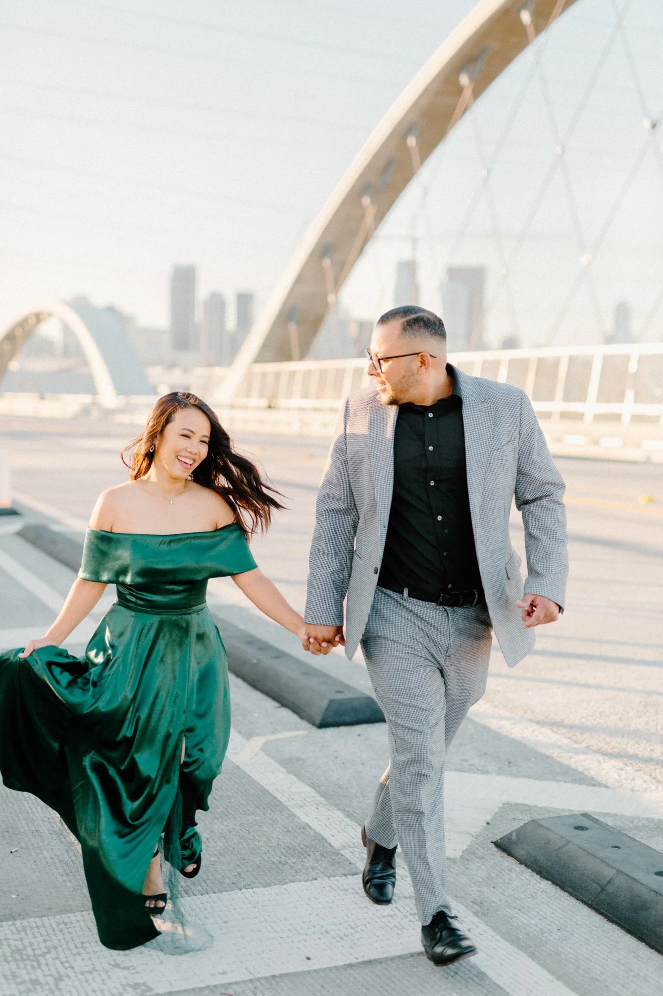 Engaged couple dressed in a glamorous emerald green dress and stylish suit run down 6th street bridge in downtown los angeles at sunset