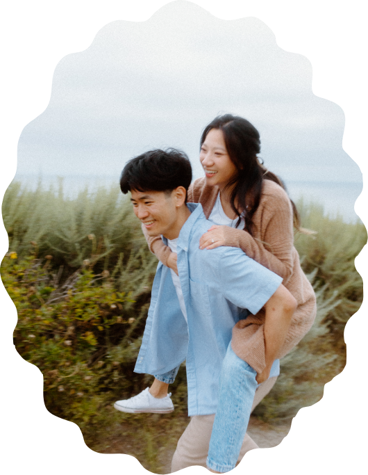 candid photography of A young couple outdoors, the woman riding on the man's back, both smiling, with greenery and a cloudy sky in the background.