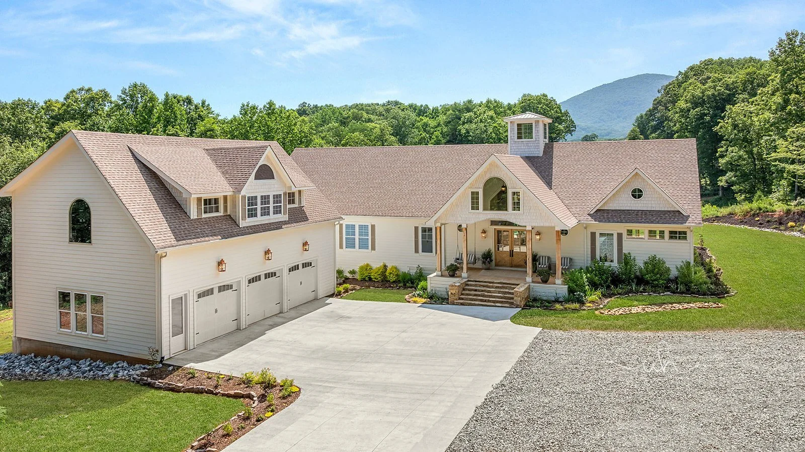 A large white house with a three-car garage and a stone staircase leading to the front porch, surrounded by green grass and trees in the background, with mountains in the distance under a blue sky.