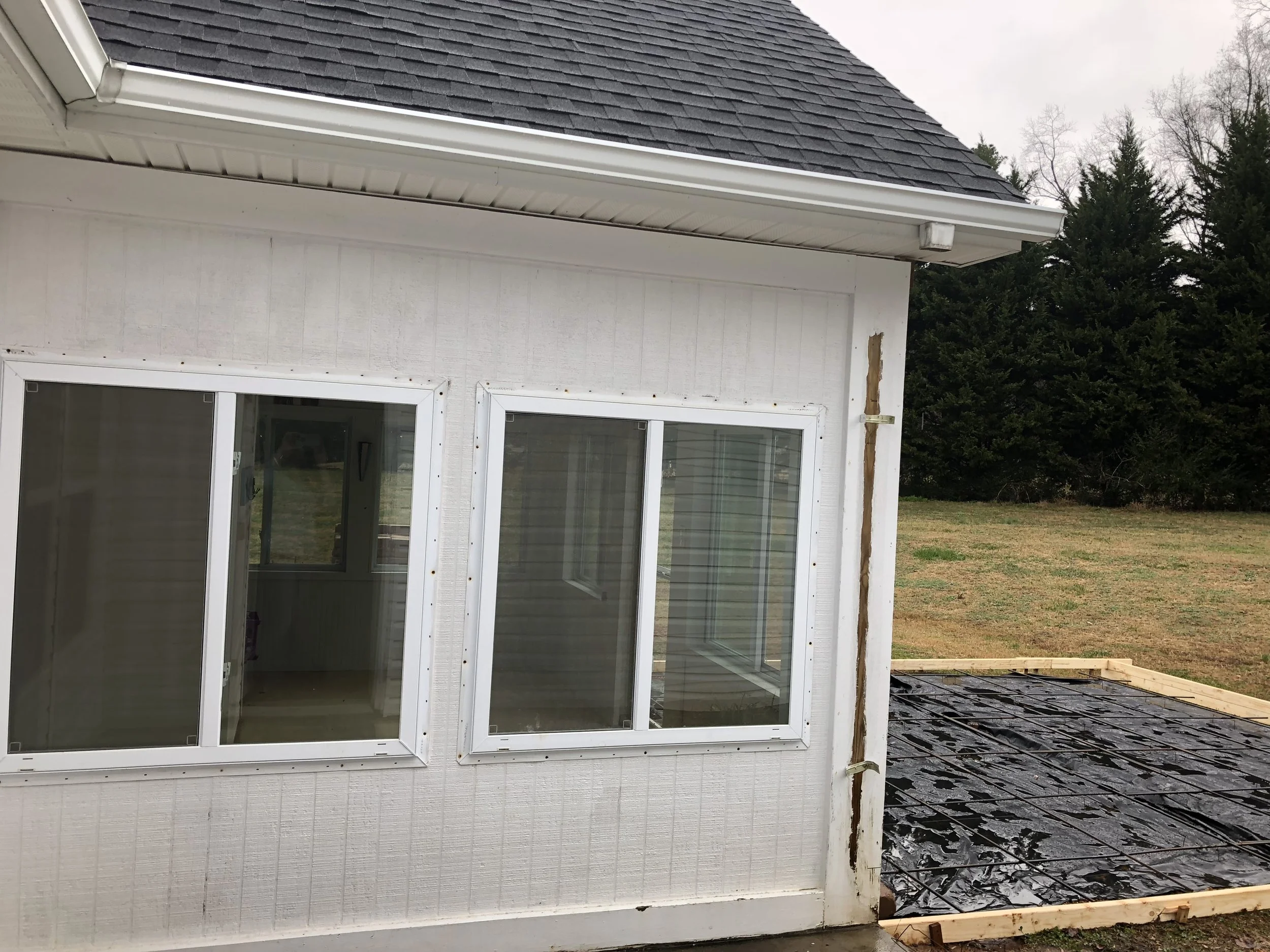 Exterior view of a house under construction with vinyl siding, large sliding glass door, and a roof covered with black shingles. The construction area has exposed framing and black waterproofing material on the ground. Trees are visible in the background.