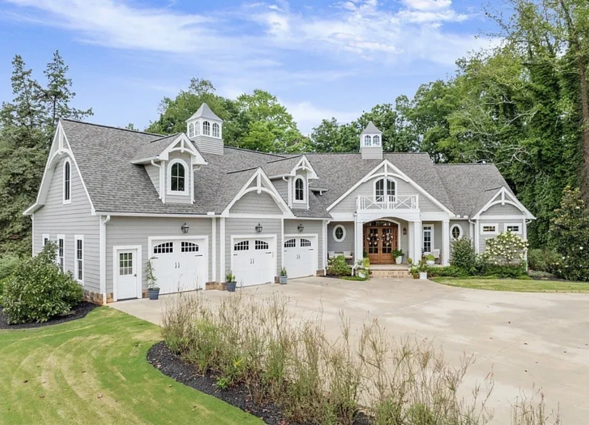 Large, gray, two-story house with multiple gables, dormer windows, and a front porch with wooden double doors. The house has a three-car garage and is surrounded by a well-maintained lawn and trees.