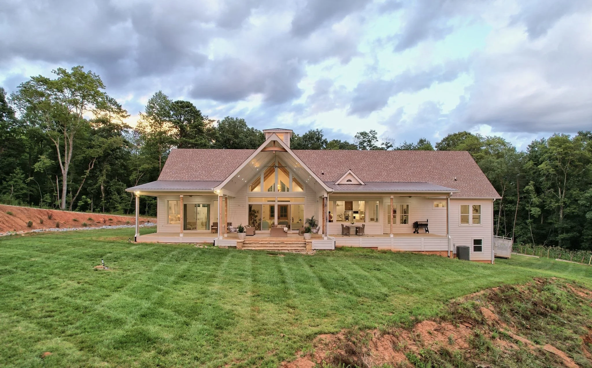 A large, modern house with a back porch and a well-maintained lawn, surrounded by trees under a cloudy sky.