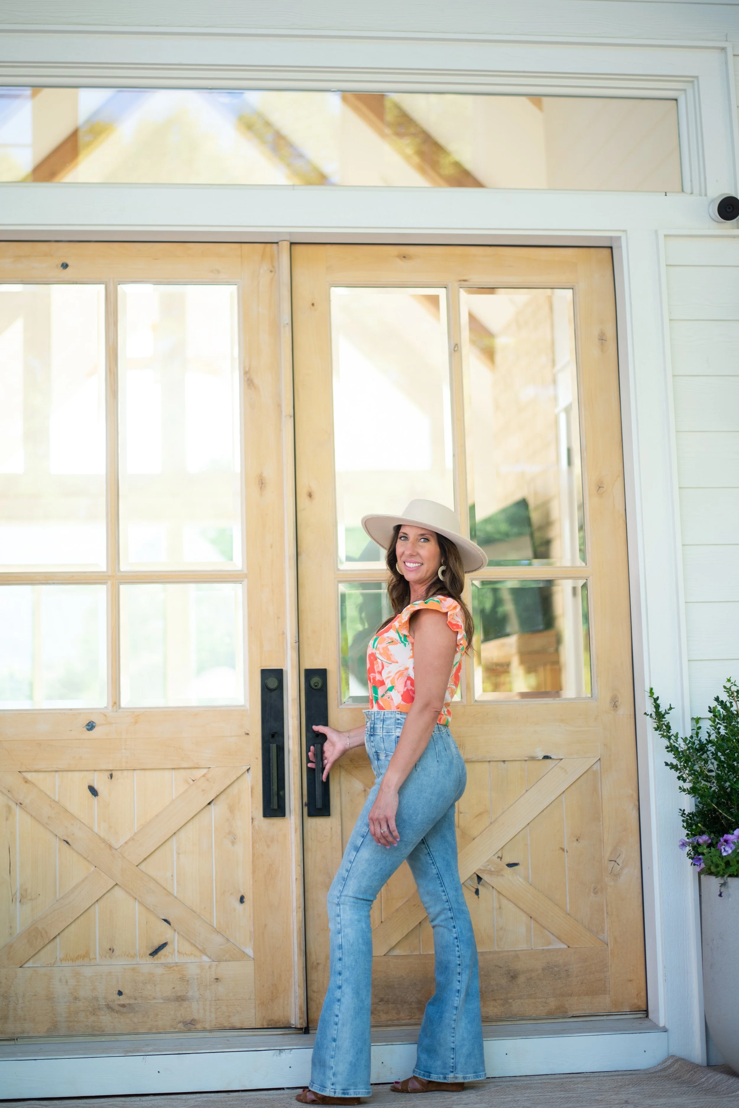 A woman in a floral top and jeans is standing by a wooden door with glass panels, holding the door handle and smiling.