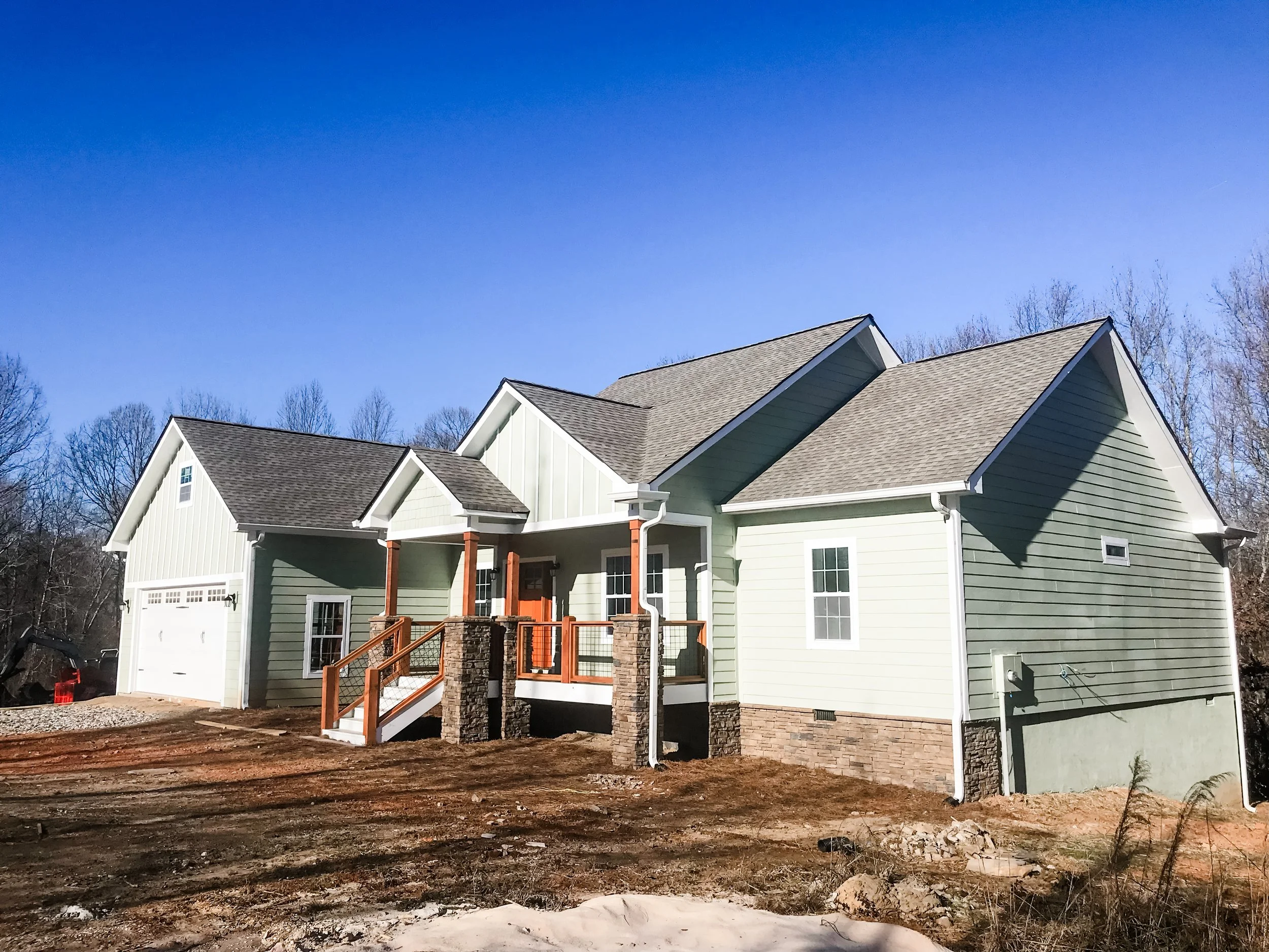 Newly built two-story house with green exterior, gray shingled roof, front porch with wooden stairs and stone pillars, surrounded by dirt and construction site.