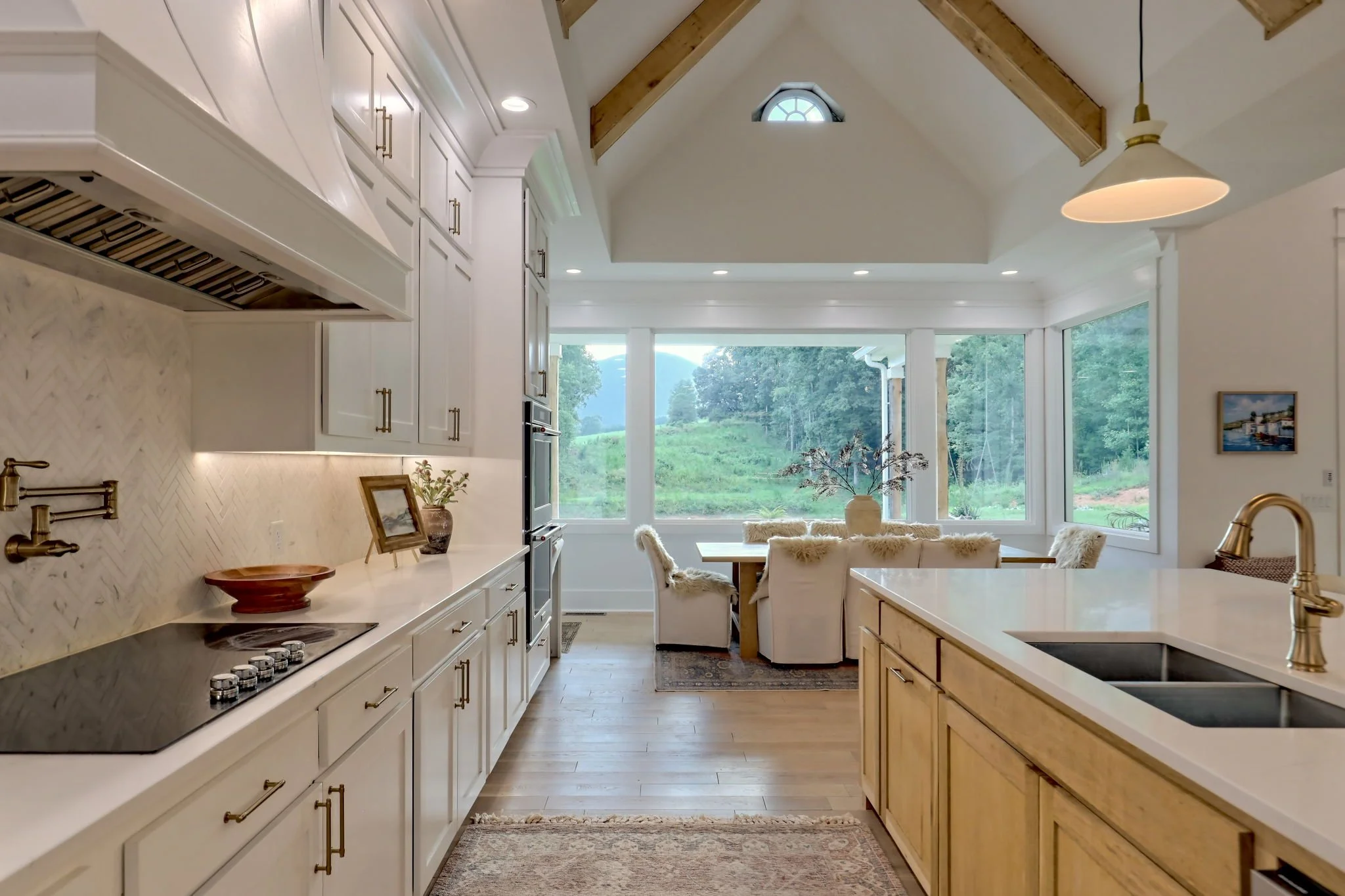 Kitchen with white cabinets, a marble backsplash, a view of large windows showing green hills outside, a dining area with plush chairs, ceiling lights, and a vaulted ceiling with wooden beams.