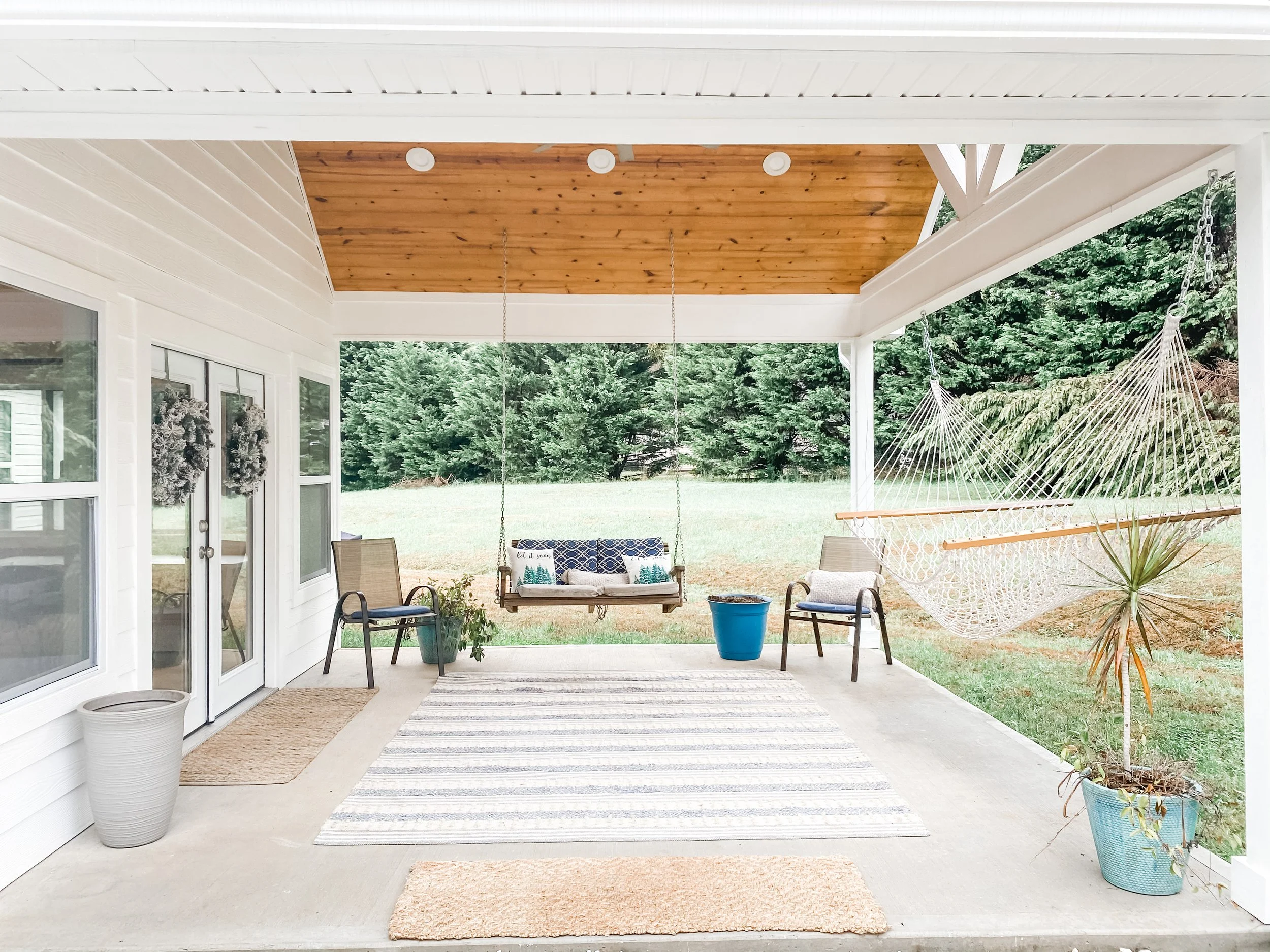 Covered patio with outdoor furniture, a hammock, potted plants, and a grassy yard with trees in the background.