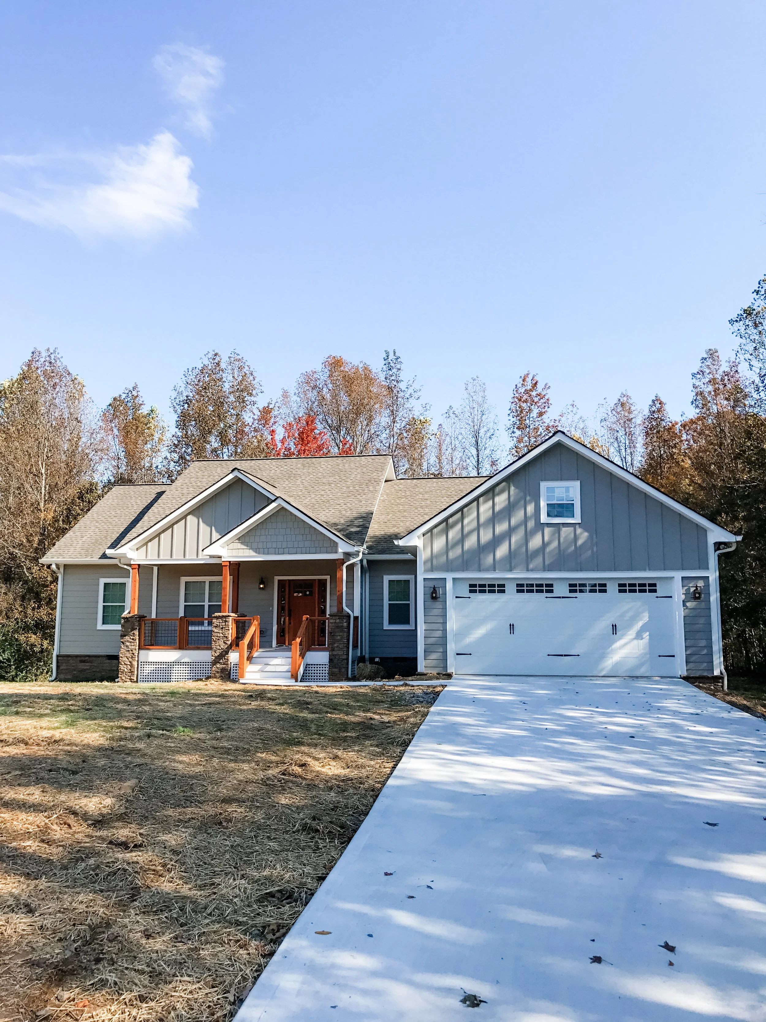 New house with a driveway, two-car garage, porch, front steps, and surrounded by trees with fall foliage.
