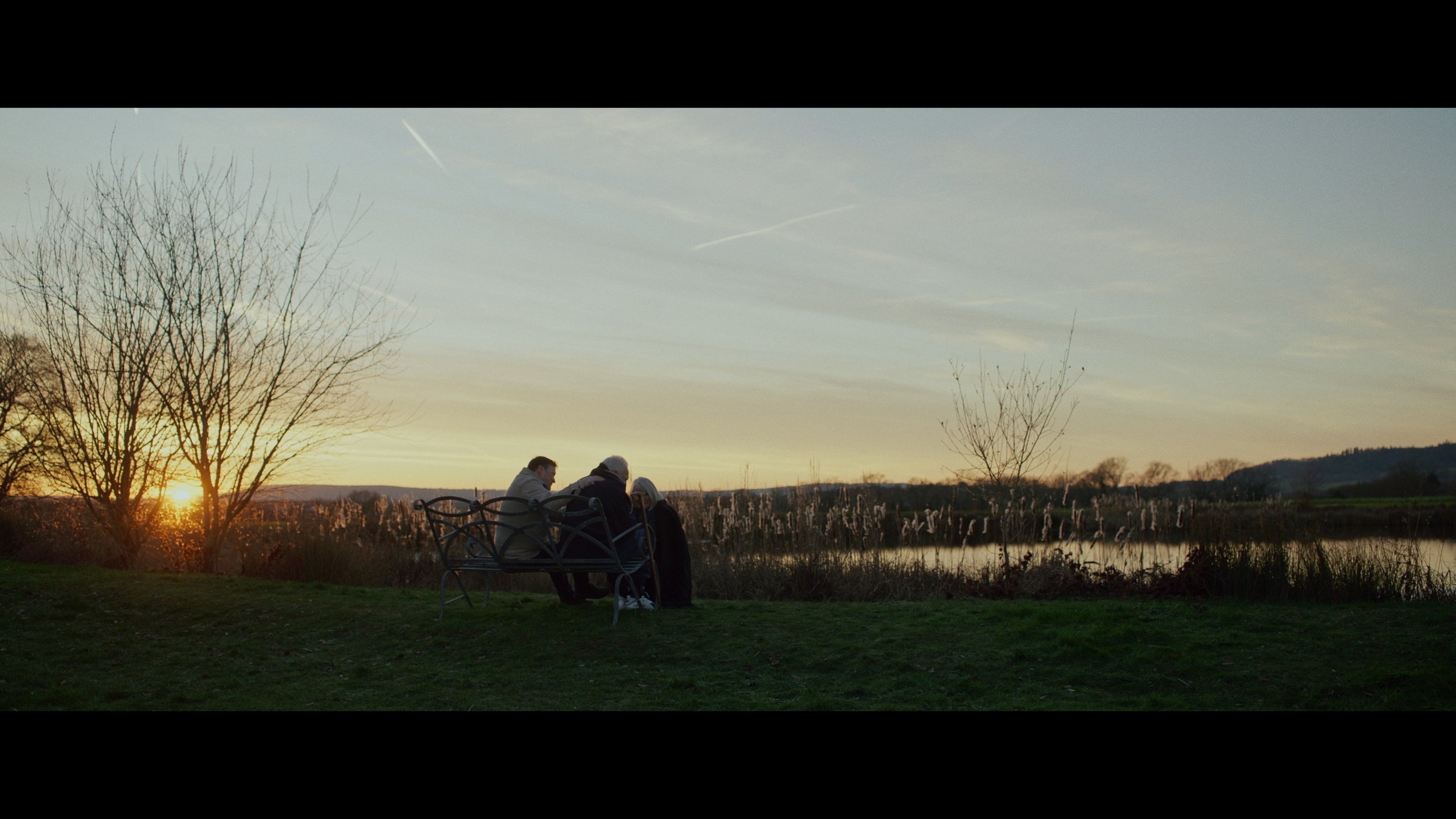 A group of people sitting on a bench by a lake during sunset, surrounded by bare trees and tall grass.