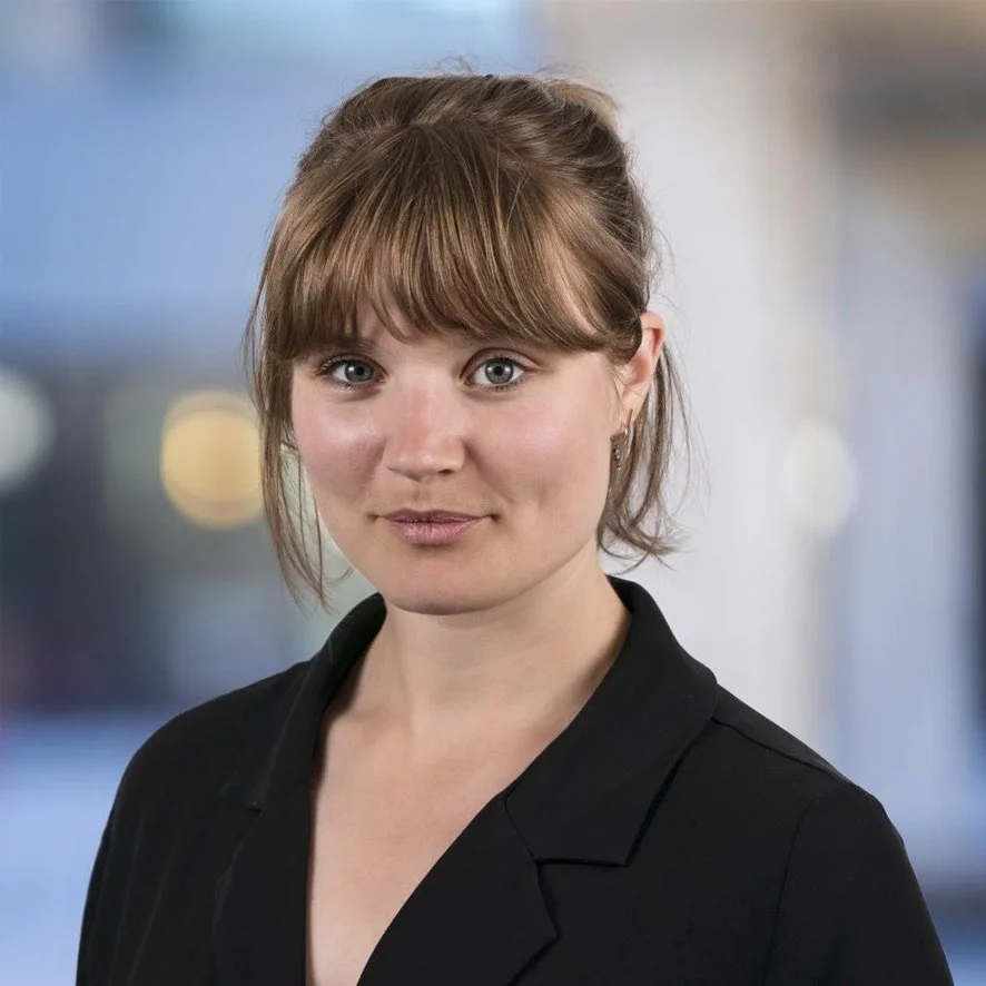 A woman with brown hair styled in a loose updo and straight bangs, wearing a black blazer, standing against a blurred background.