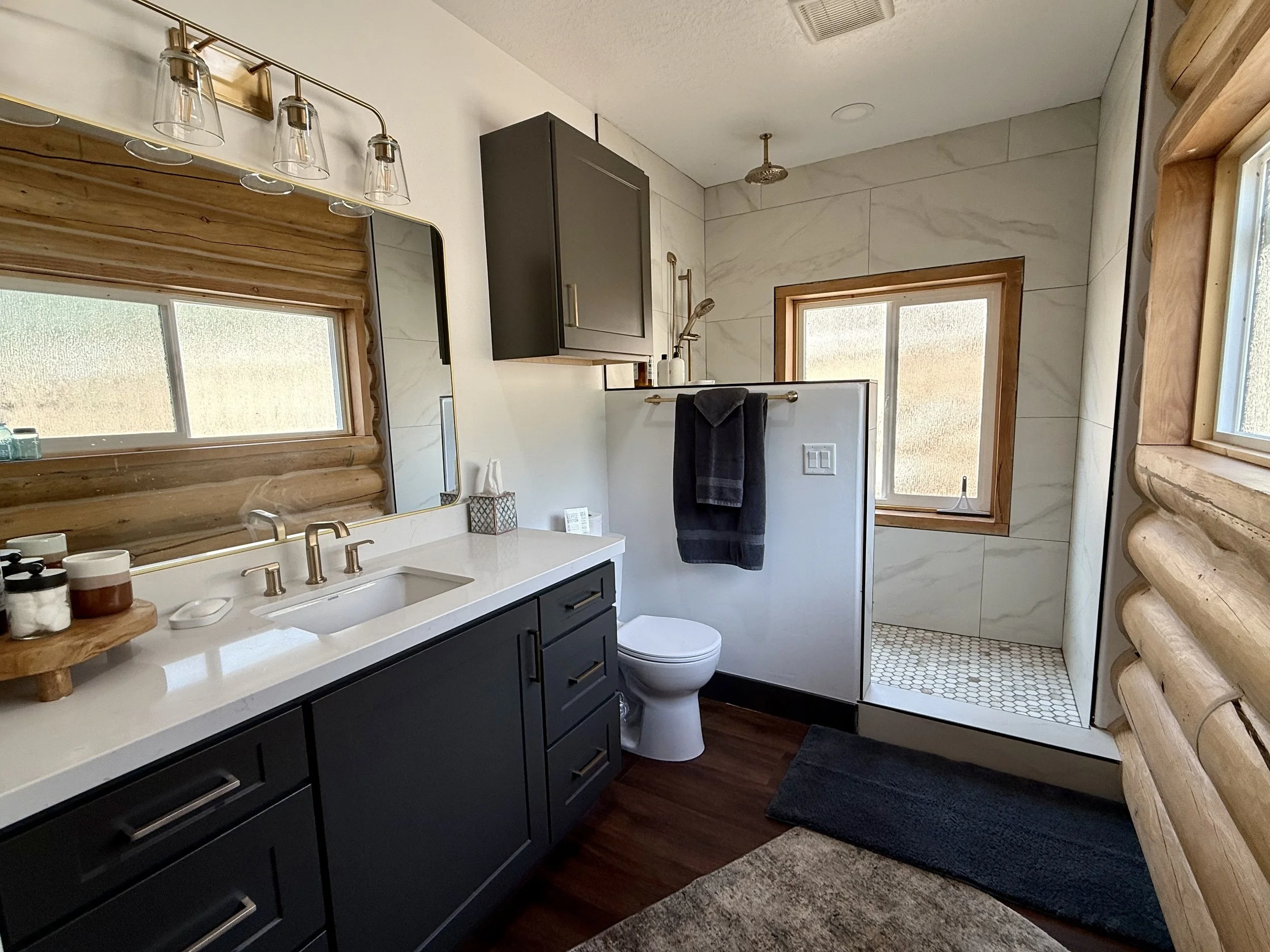 A modern bathroom featuring a dark blue vanity with a white countertop, a large mirror, a window with wooden trim, a walk-in shower with wooden framing and white hexagonal floor tiles, a toilet with a black towel hanging nearby, and wooden log cabin walls.