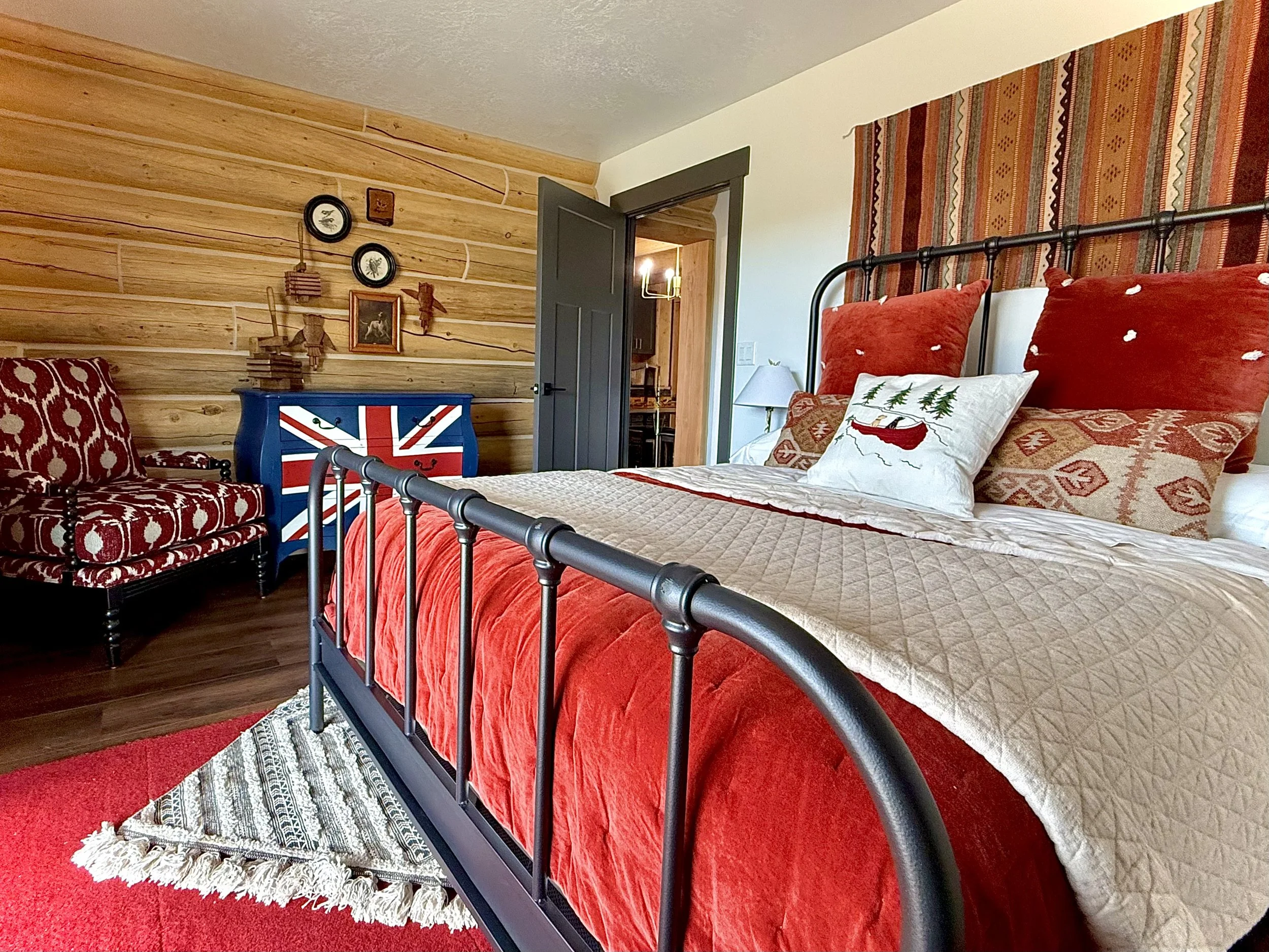 A bedroom decorated with a Christmas theme featuring a black metal bed frame, red and white pillows with a reindeer and trees, a quilted beige blanket, and a red bedspread. There are wooden walls, a colorful armchair with a red and white pattern, a b