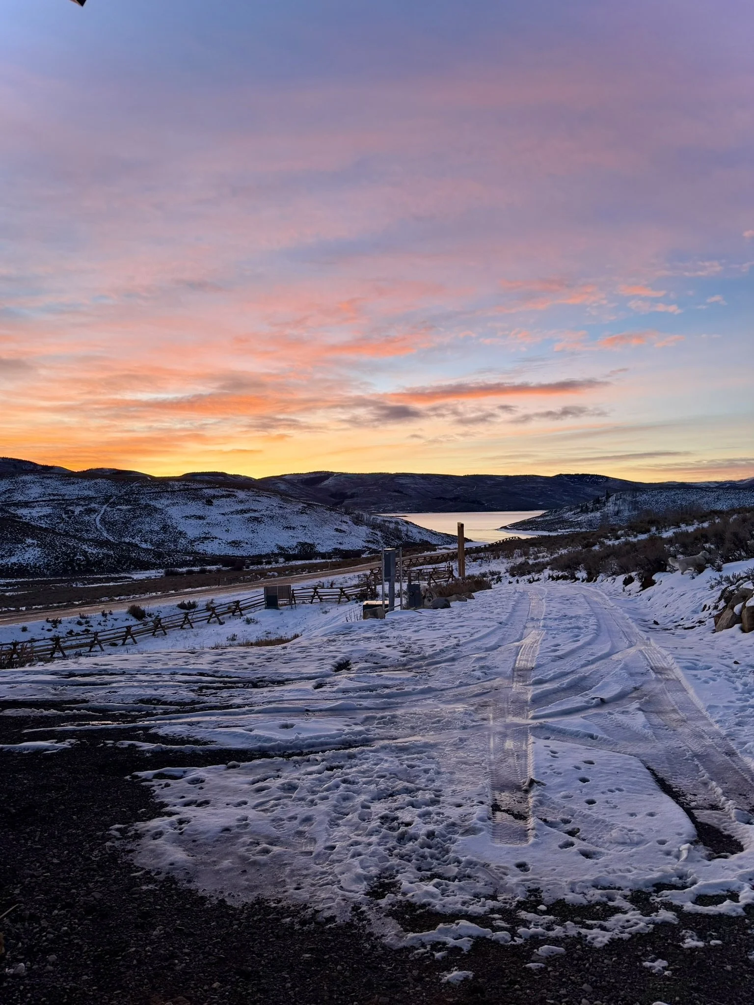 Snow-covered dirt path leading towards a lake at sunset, surrounded by hilly terrain with a colorful sky.