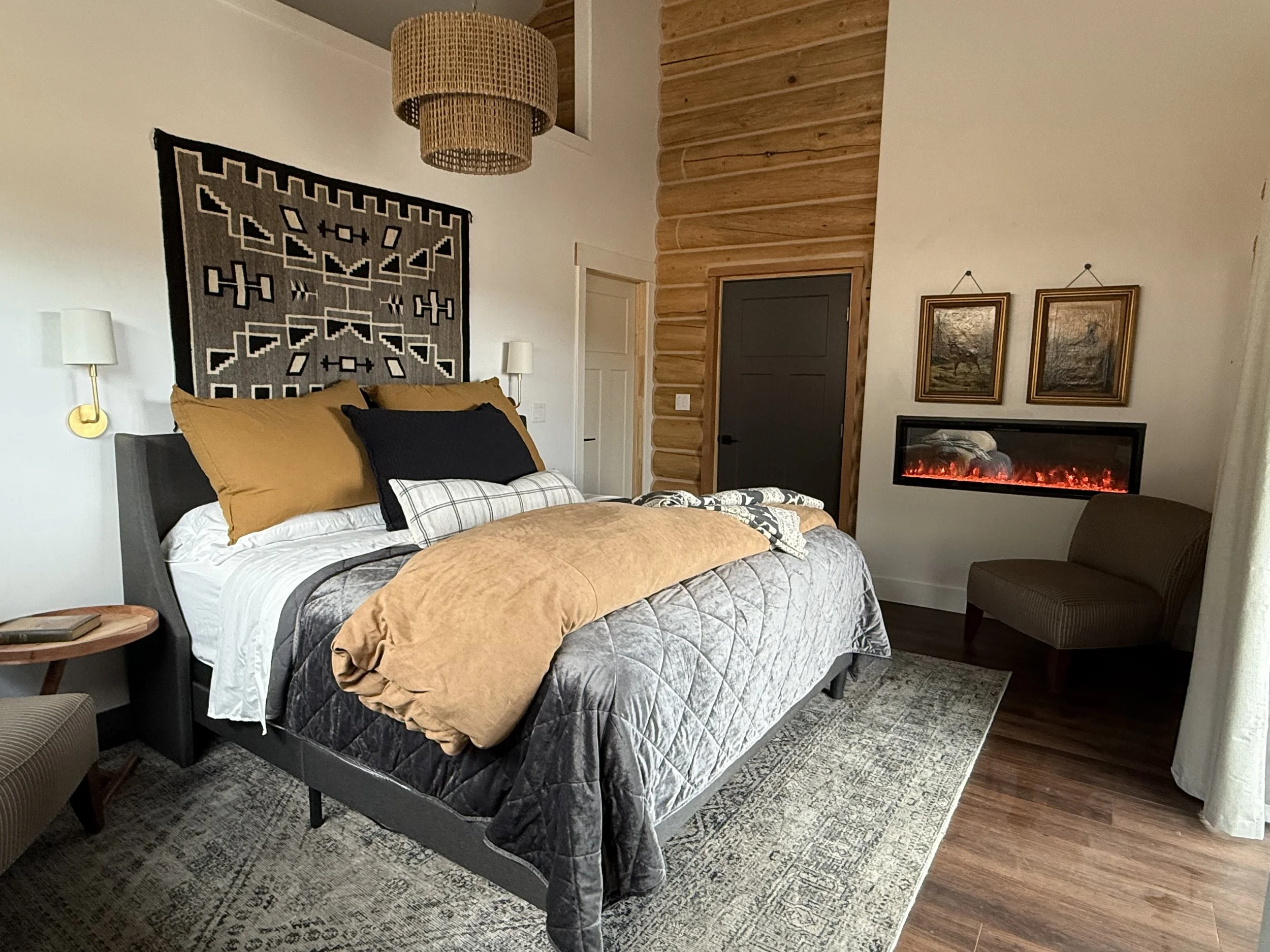 A bedroom with a bed covered in gray, black, white, and tan bedding, featuring multiple pillows, and a patterned tapestry hanging above it. There's a woven ceiling lamp, wall-mounted lamps on either side of the bed, a small wooden side table with books, a patterned area rug, a brown upholstered chair, dark wood flooring, a wall with wood paneling, a modern electric fireplace, and two framed landscape paintings.