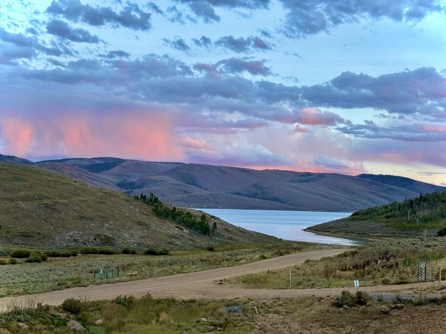 Scenic landscape with rolling hills, a calm lake, a dirt road, scattered bushes, and a colorful sky with pink and blue clouds.