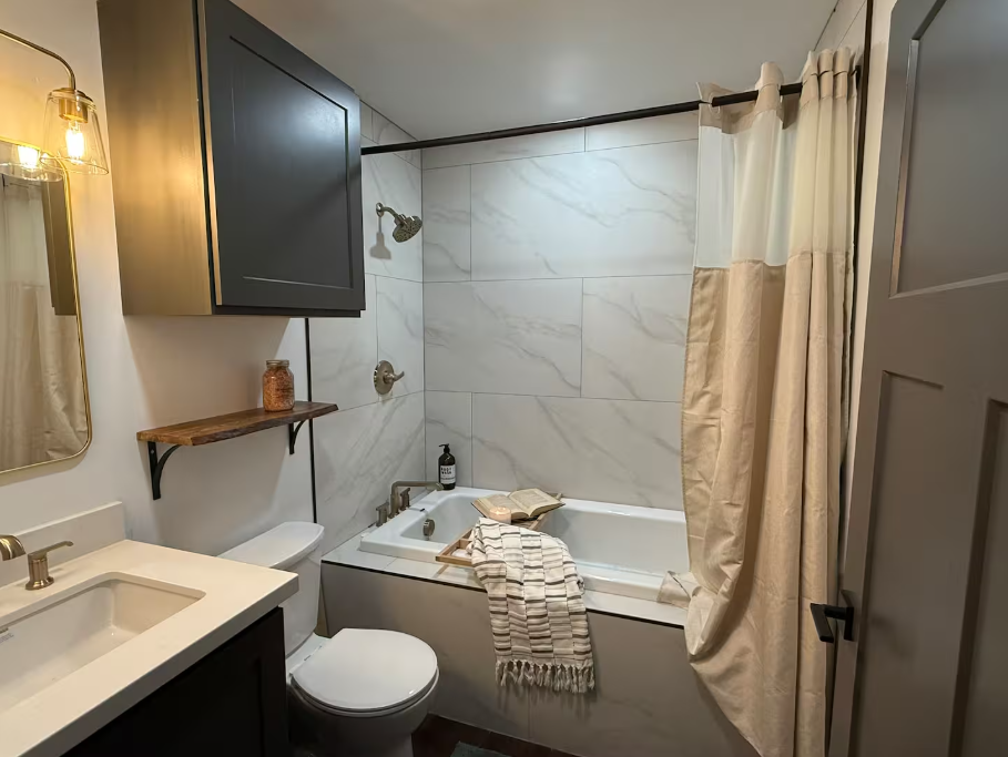 Small modern bathroom with a white bathtub, wooden shelf, gray cabinet, and beige shower curtain.