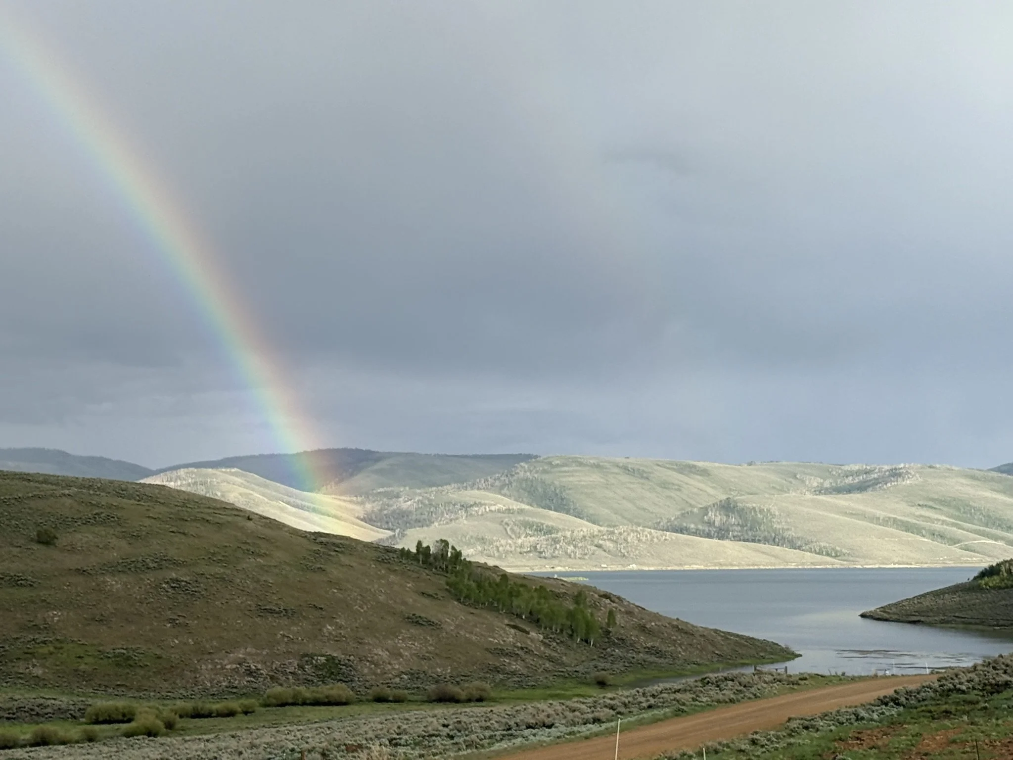 A landscape with a rainbow over rolling hills and a body of water, cloudy sky.