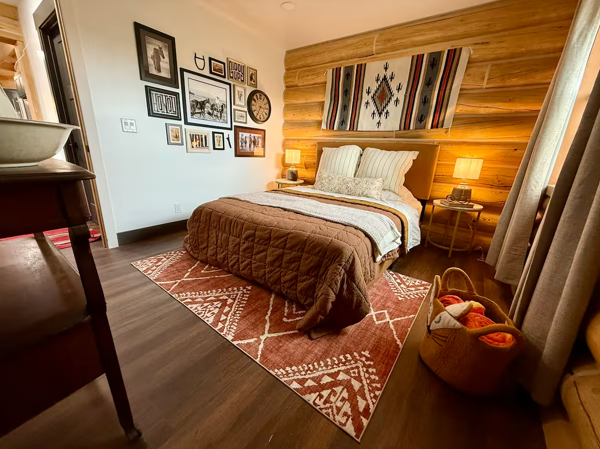 Cozy bedroom with a log cabin style wooden accent wall behind the bed, framed pictures and artwork on the adjacent wall, and warm lighting from lantern-style table lamps. The bed has striped and floral pillows, and a southwestern-style tapestry hangs