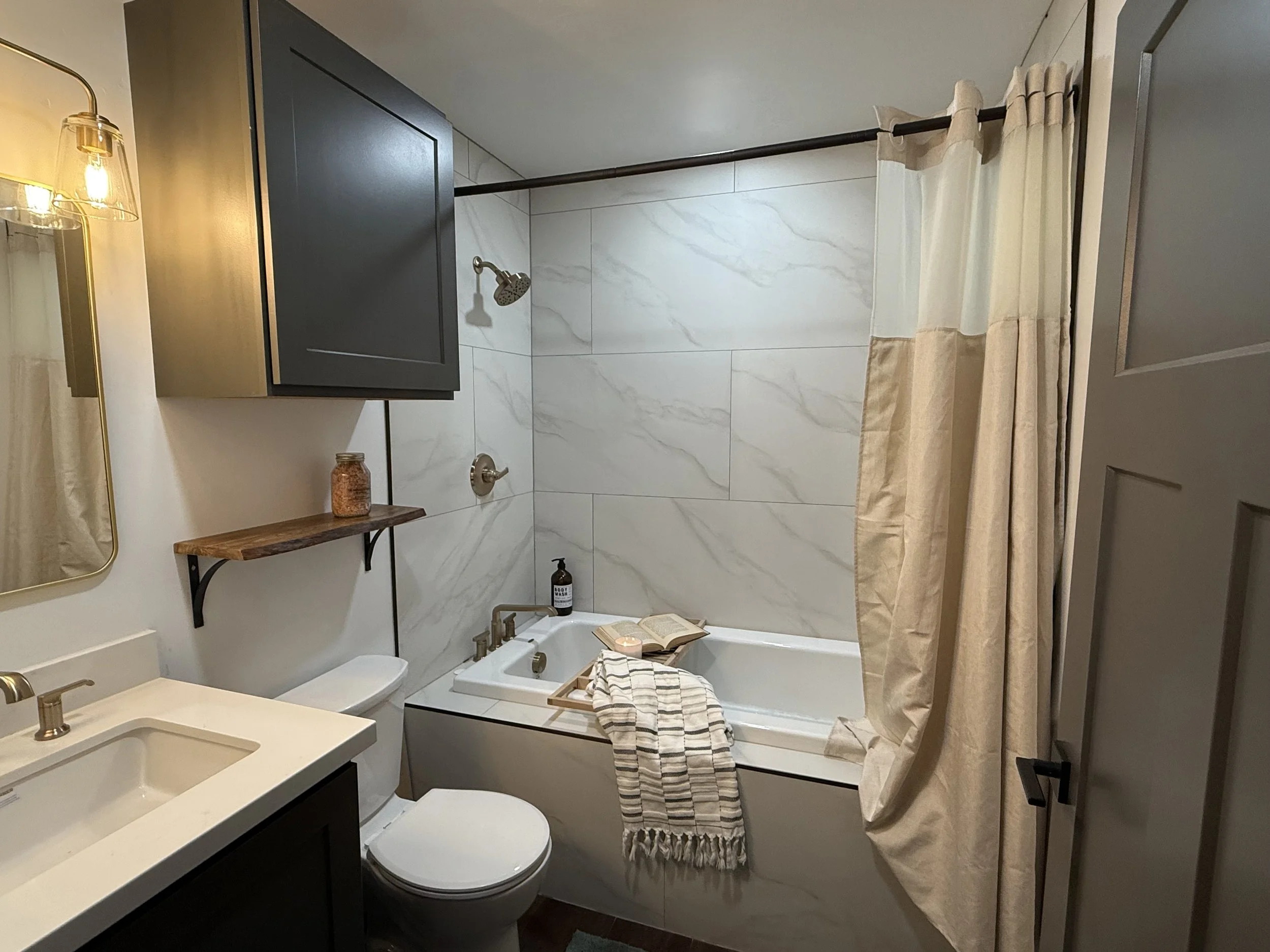 A modern bathroom with a bathtub, showerhead, beige curtain, dark wall cabinet, white sink with black cabinet, mirror, small wooden shelf, and towel, soap, and book on the tub.