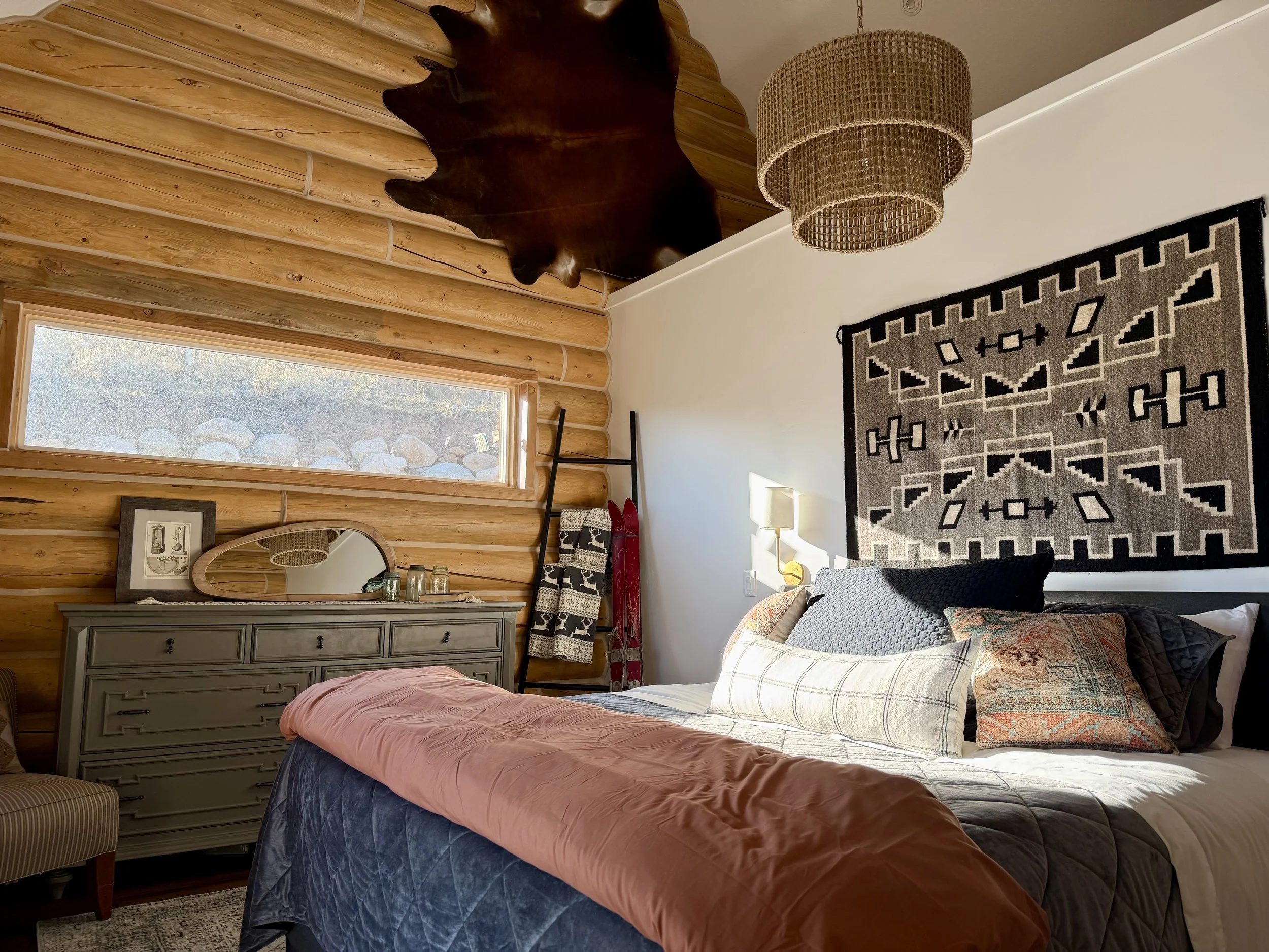 A cozy bedroom with wooden log walls, a large bed with multiple pillows, a black and white geometric wall hanging, a rustic dresser with a mirror, a window letting in natural light, and a woven hanging lamp.
