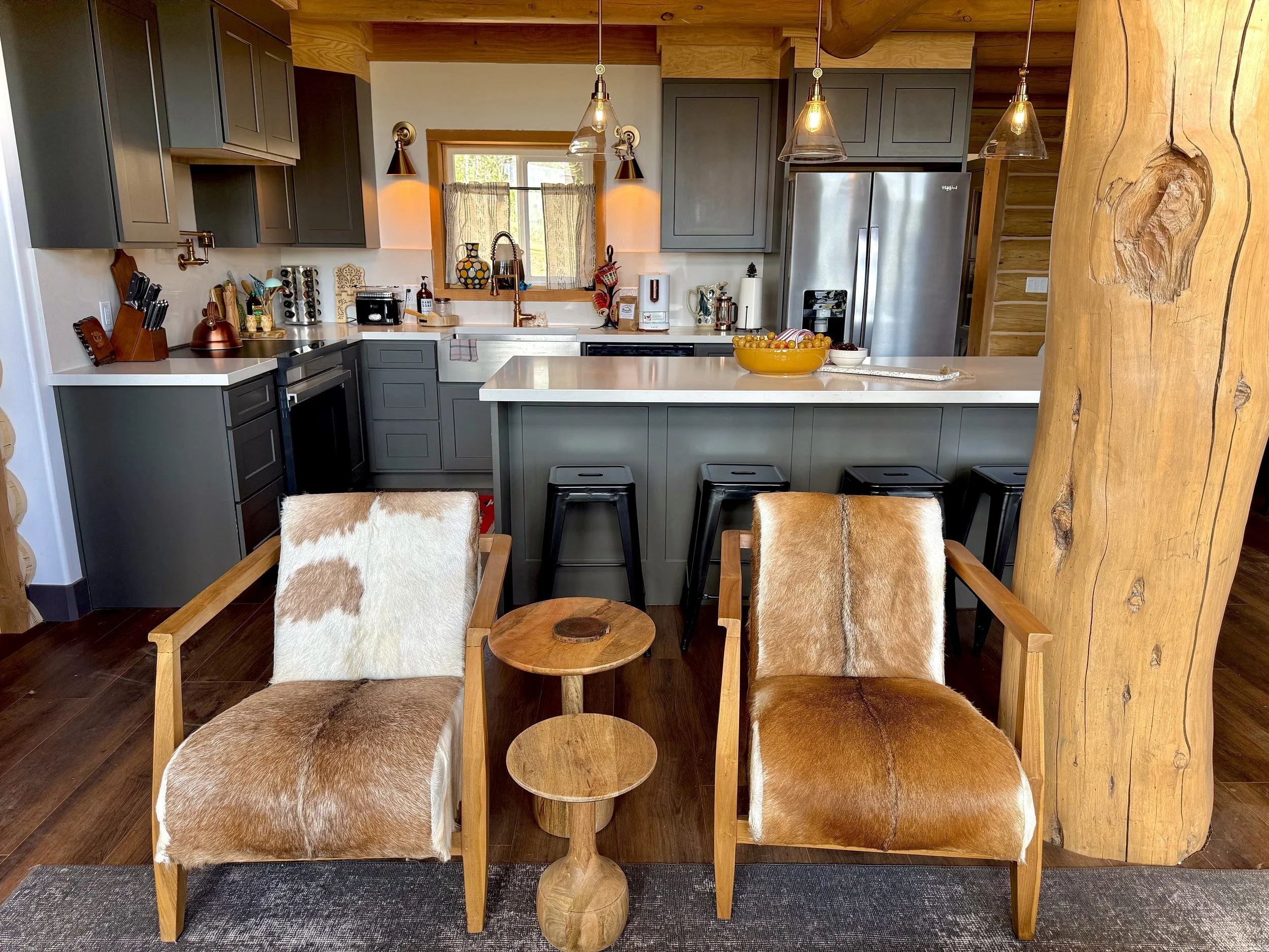 A cozy kitchen and sitting area with two wooden chairs with cowhide upholstery, a small wooden side table, and modern grey cabinets with stainless steel appliances, and a breakfast bar with four black stools.