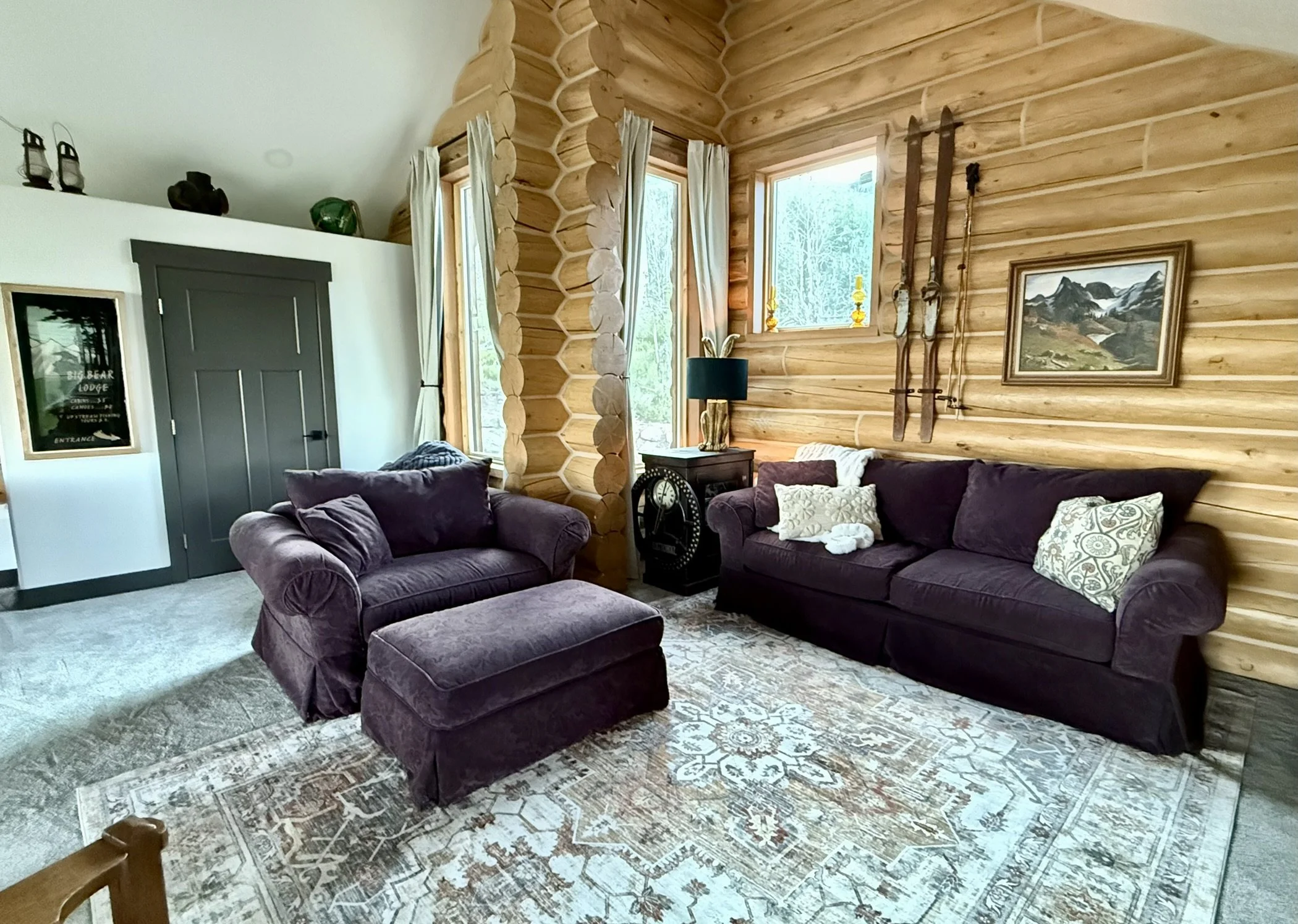 Living room with wood-paneled walls, a dark purple sofa with patterned pillows, a matching armchair with an ottoman, a rug, and a small side table with a lamp. There are tall windows with curtains, and decorative winter skis and a painting on the wal