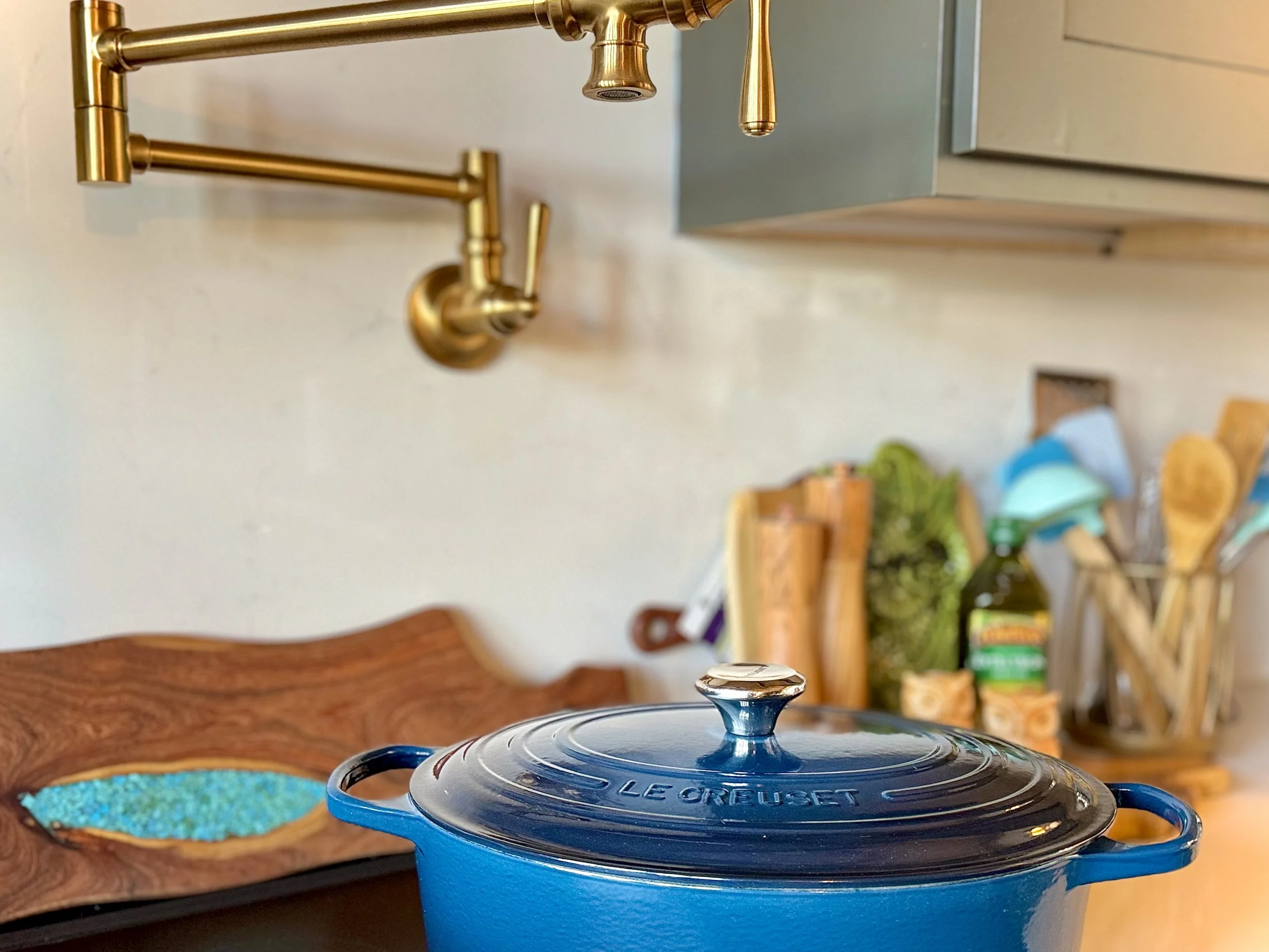 A blue Le Creuset cast iron Dutch oven with a lid is on a kitchen countertop. Above it, there is a gold-colored kitchen faucet. In the background, there are kitchen utensils, a cutting board, a green leafy vegetable, a bottle, and a utensil holder on