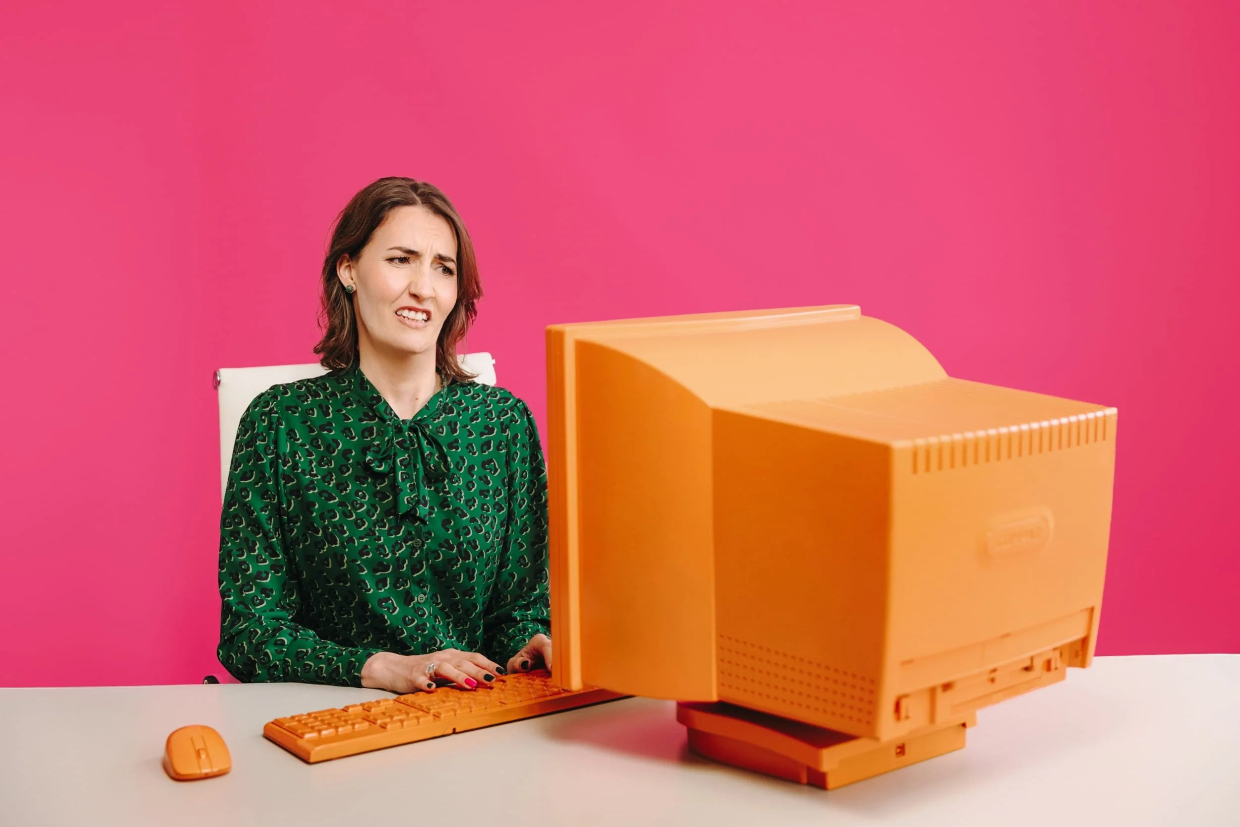 Portretfoto van freelance SaaS-copywriter en contentspecialist Marije van Wijngaarden, gekleed in een groene blouse met zwarte panterprint, fronsend en werkend aan een oranje vintage computermonitor met oranje toetsenbord en muis.