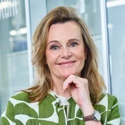 Portret van een vrouw met lichtbruin haar en een groene en witte blouse, voor een glazen achtergrond.