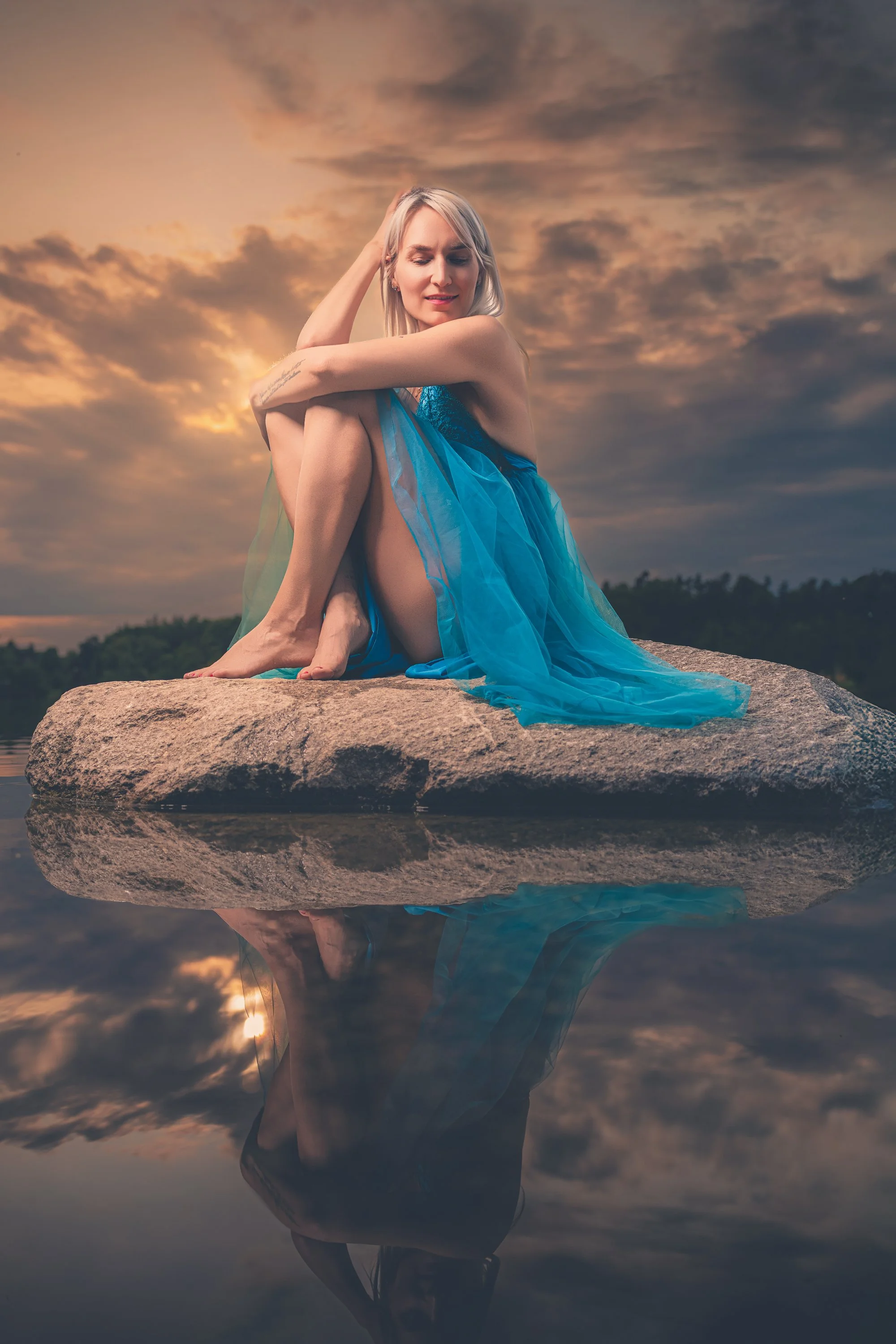 A woman in a flowing blue dress sitting on a large rock by the water at sunset, with her reflection visible in the water.