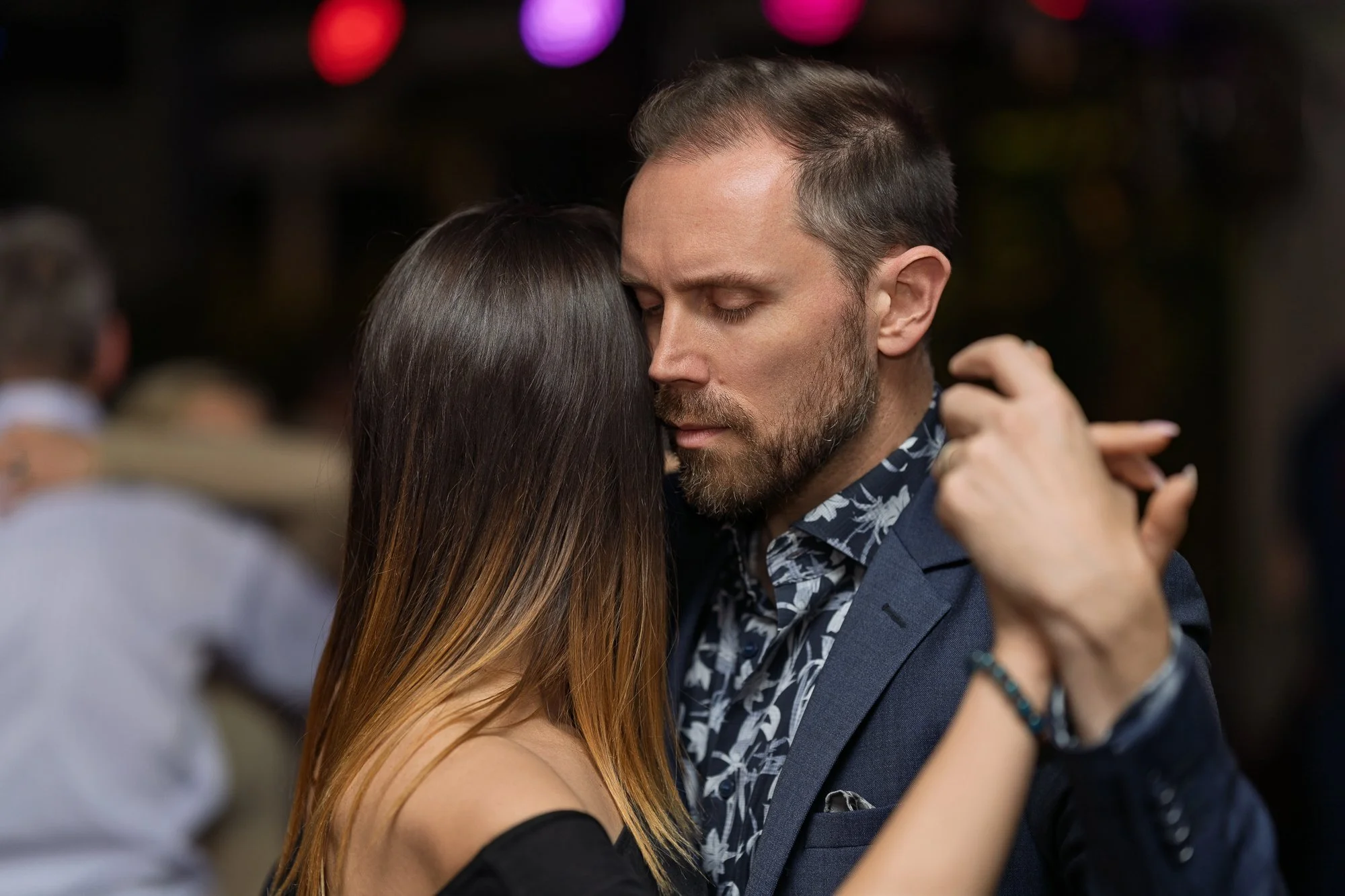 A couple dancing closely at a social event, with their foreheads touching and eyes closed, in a dimly lit setting.
