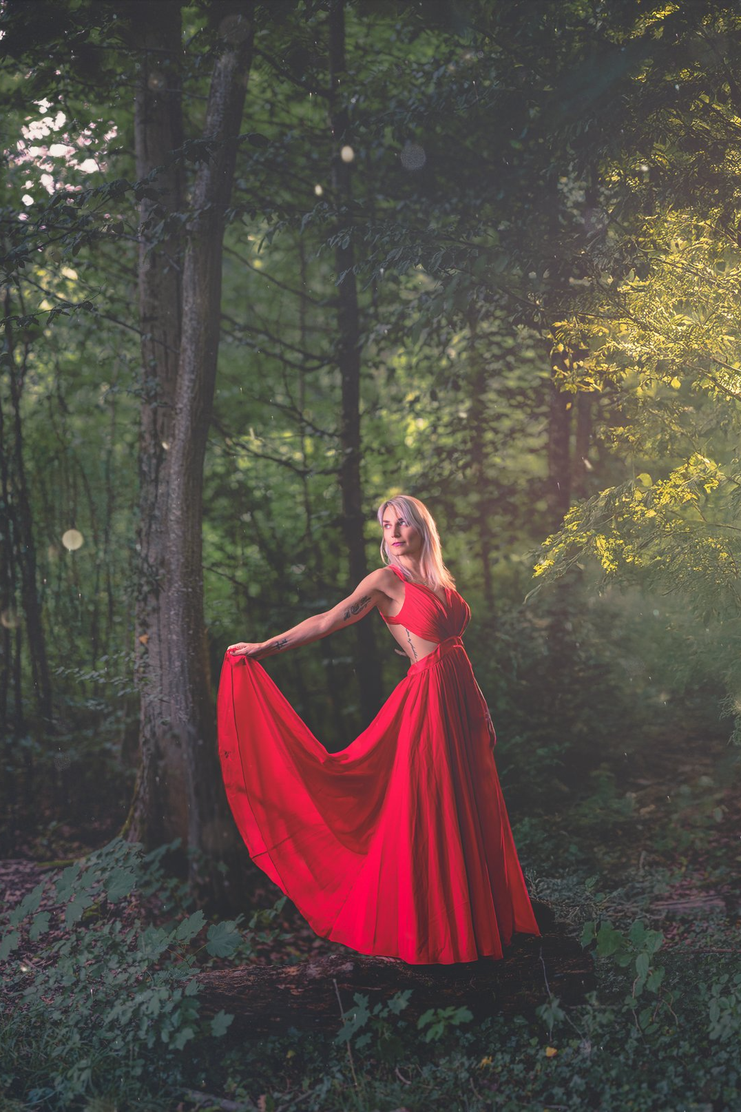 A woman in a flowing red dress standing in a lush green forest, holding the dress out to the side with one arm, while sunlight filters through the trees.